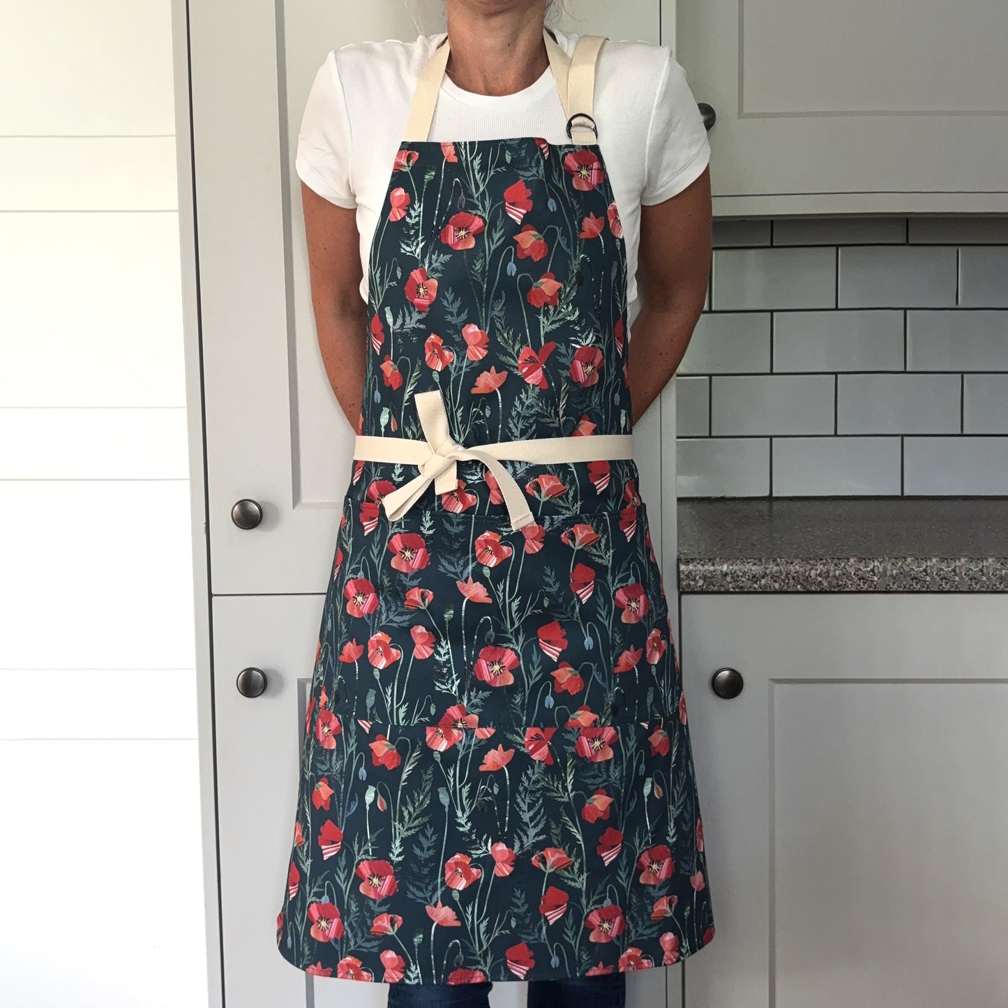 A lady is wearing the Poppy apron with two pockets on the front and an adjustable neck strap in a grey kitchen.  She has tied the apron around her waist.