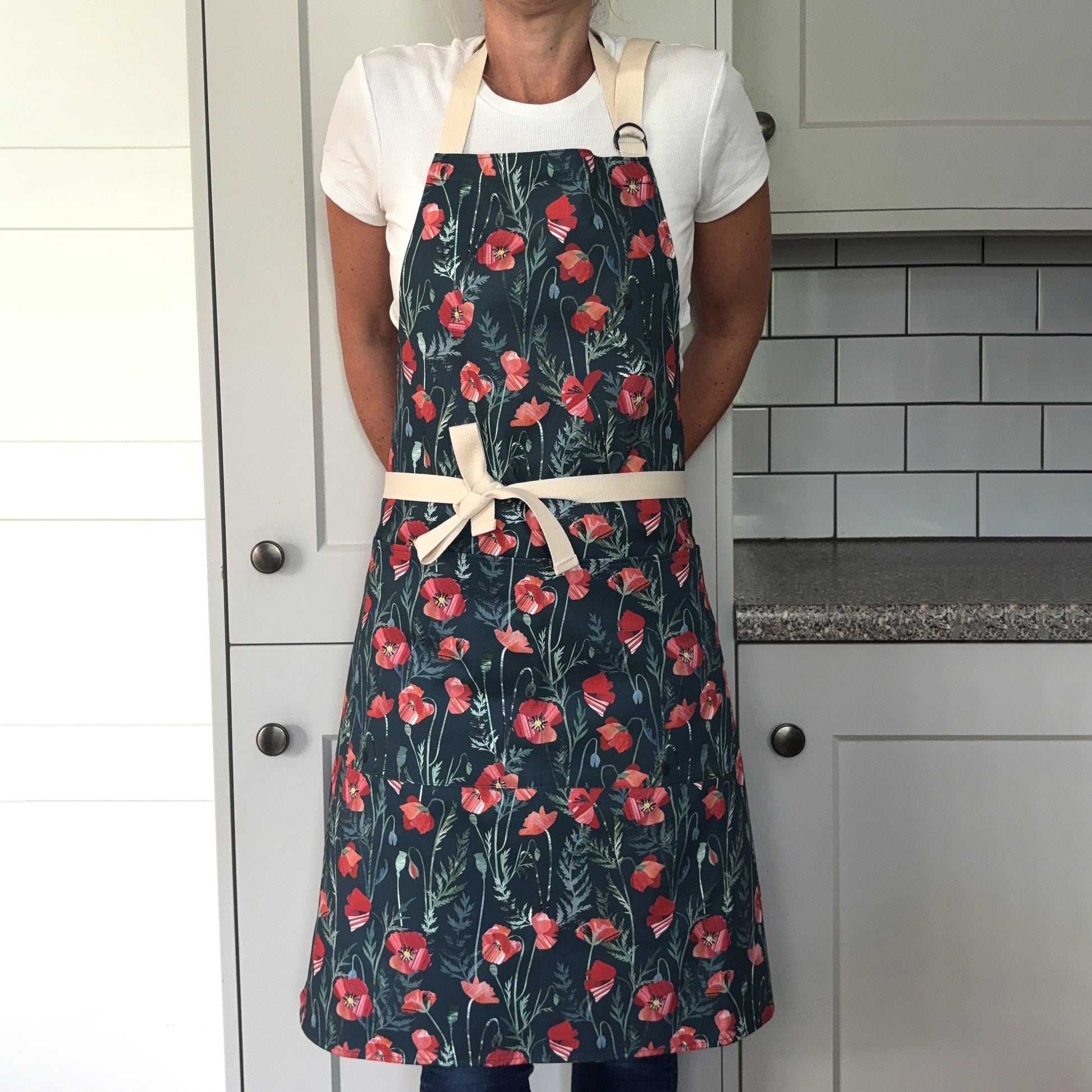 A lady is wearing the Poppy apron with two pockets on the front and an adjustable neck strap in a grey kitchen.  She has tied the apron around her waist.