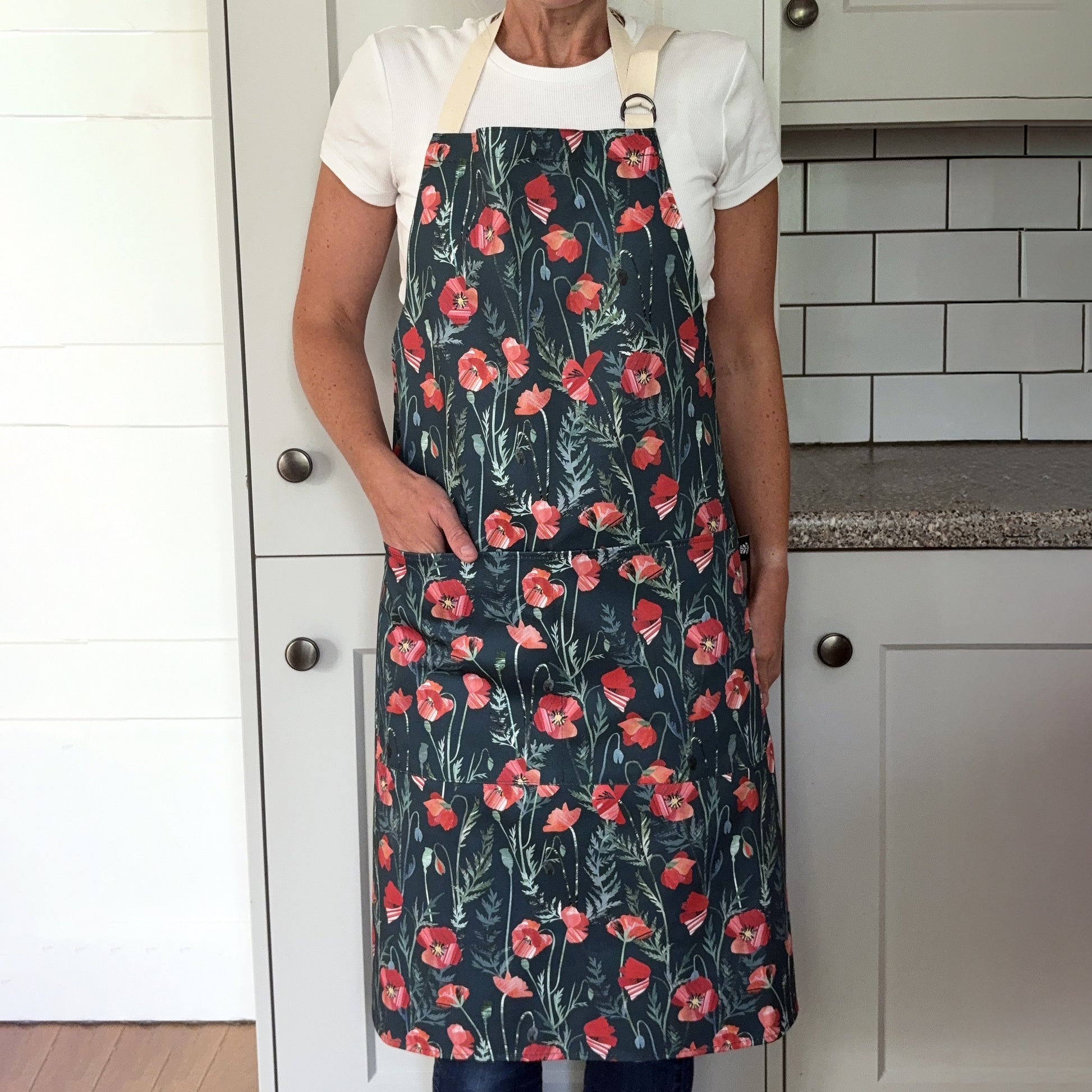 A lady is wearing the Poppy apron with two pockets on the front and an adjustable neck strap in a grey kitchen.  She has her hand in one of the front pockets.