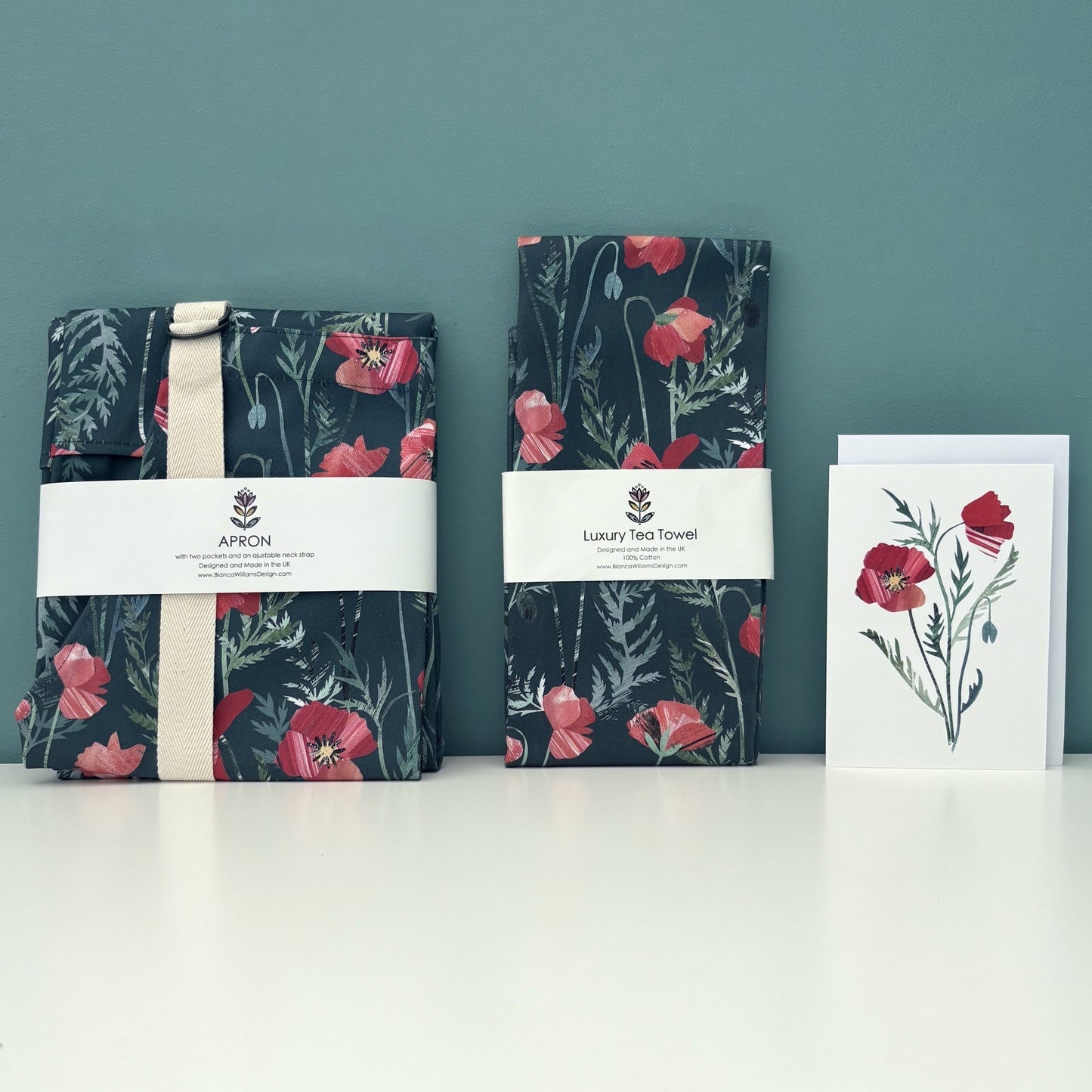 A Packaged poppy apron and matching Tea Towel with a coordinating poppy card have been placed on a white shelf against a blue green background.
