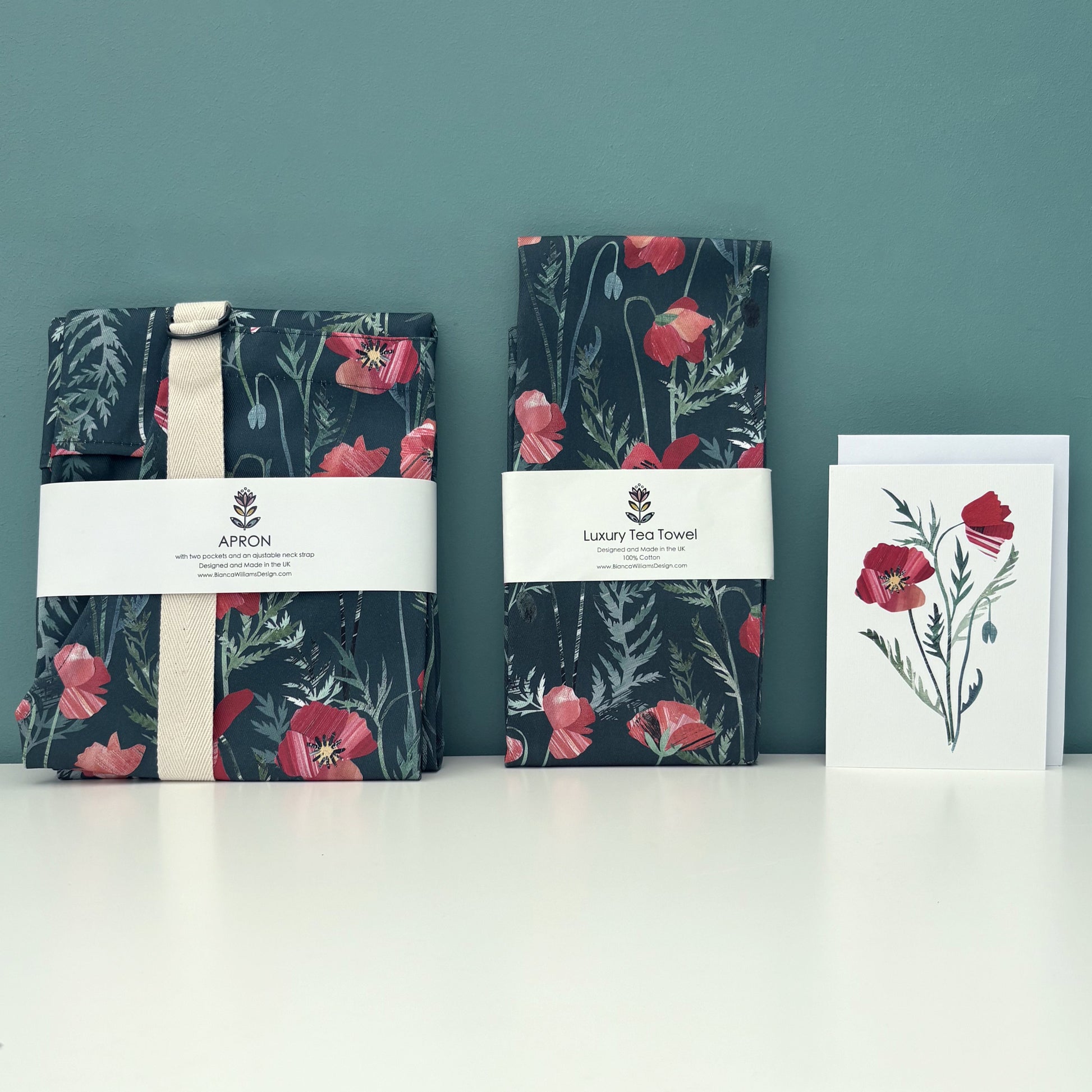 A Packaged poppy apron and matching Tea Towel with a coordinating poppy card have been placed on a white shelf against a blue green background.