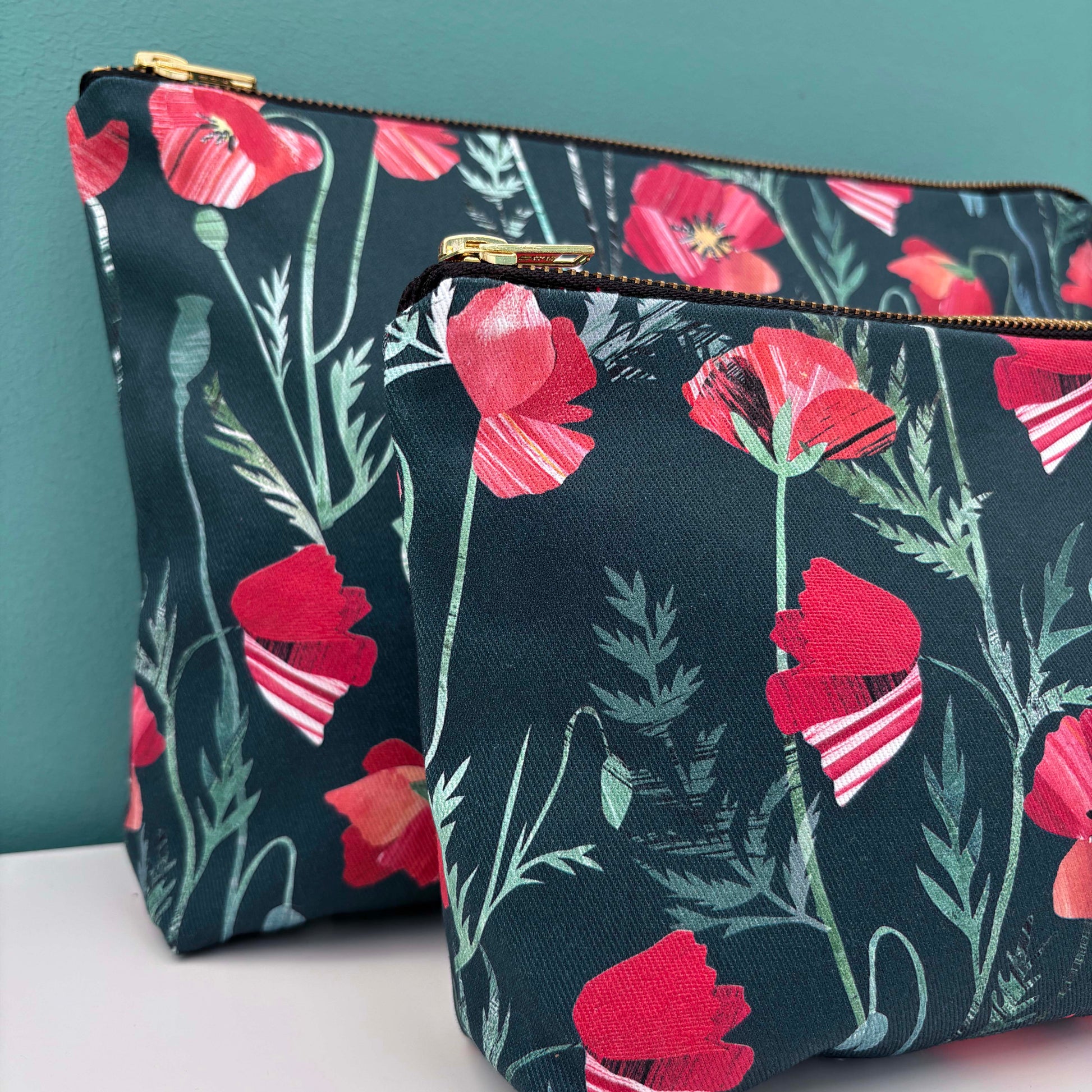 Two floral pouches one large and one smaller with red poppies on a dark background against a teal wall.