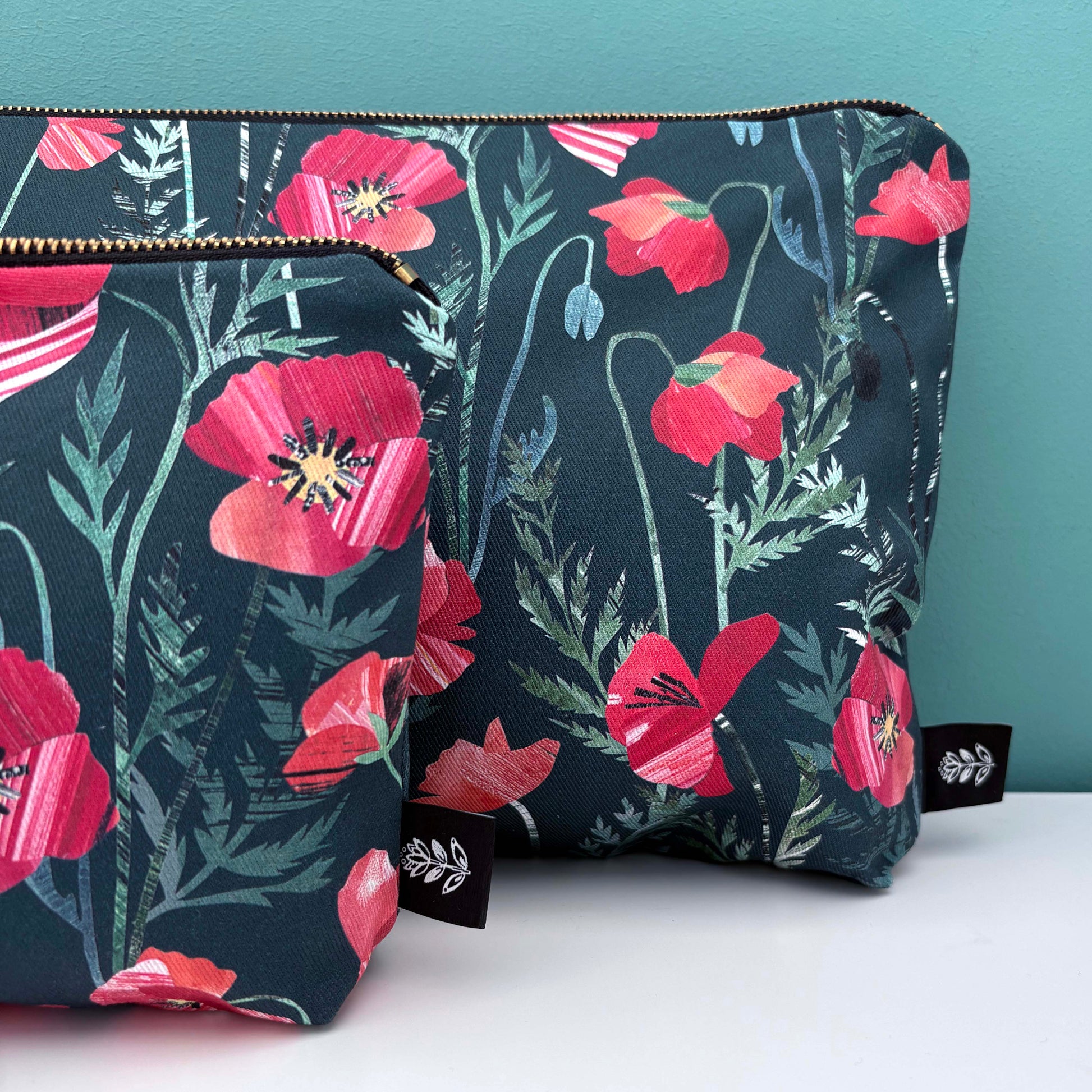A close up of the large and small poppy wash bags.  They have been placed on a white shelf against a blue green background.  The have gold metal zippers and you can see the brand label sewn into the seams.