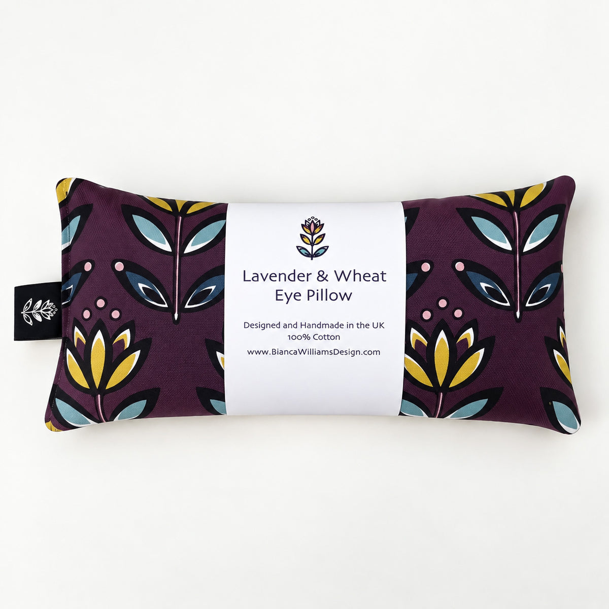 A rectangular lavender and wheat eye pillow with a dark purple background and a colorful botanical pattern in yellow, teal, and navy. A white paper label is wrapped around the centre, displaying the text 'Lavender & Wheat Eye Pillow' and the brand 'Bianca Williams Design'.