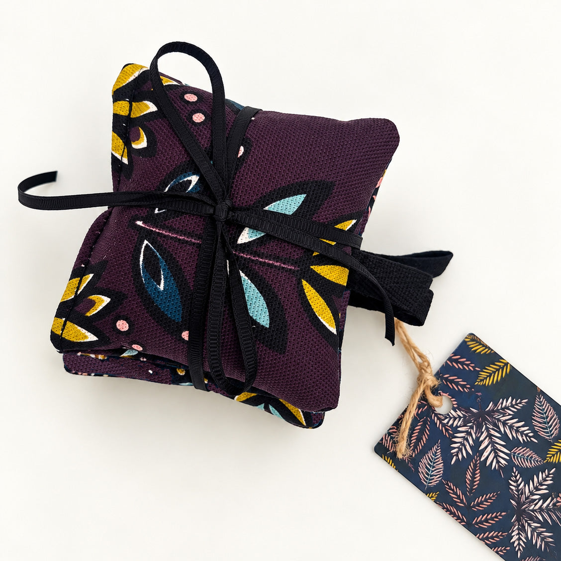 A pack of three lavender bags in a colourful abstract leaf pattern in yellow, teal, black, and white, tied with a black ribbon. A matching patterned gift tag hangs from the ribbon on a plain white background.
