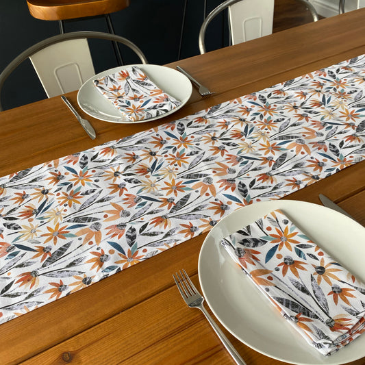 A Orange Rudbeckia Table Runner has been placed down the centre of a wooden Table.  The pattern has textured  Orange Rudbeckia flowers in different shades of orange and has grey and green leaves.  The Table has two place settings on it with matching napkins.