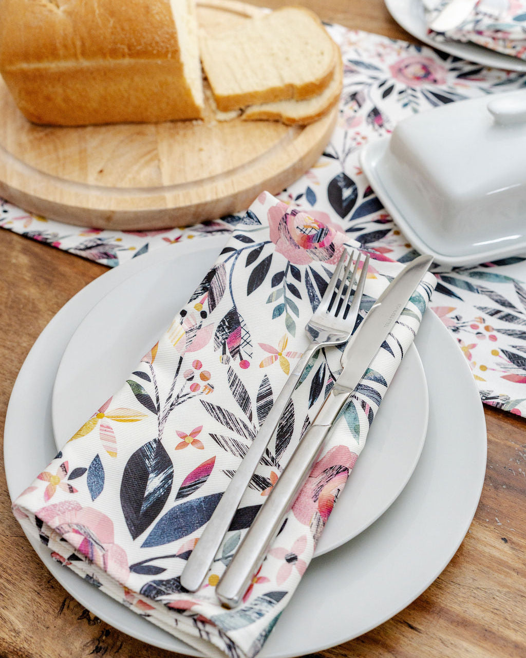 Set table with floral Roses napkin, cutlery, and bread on a wooden surface.