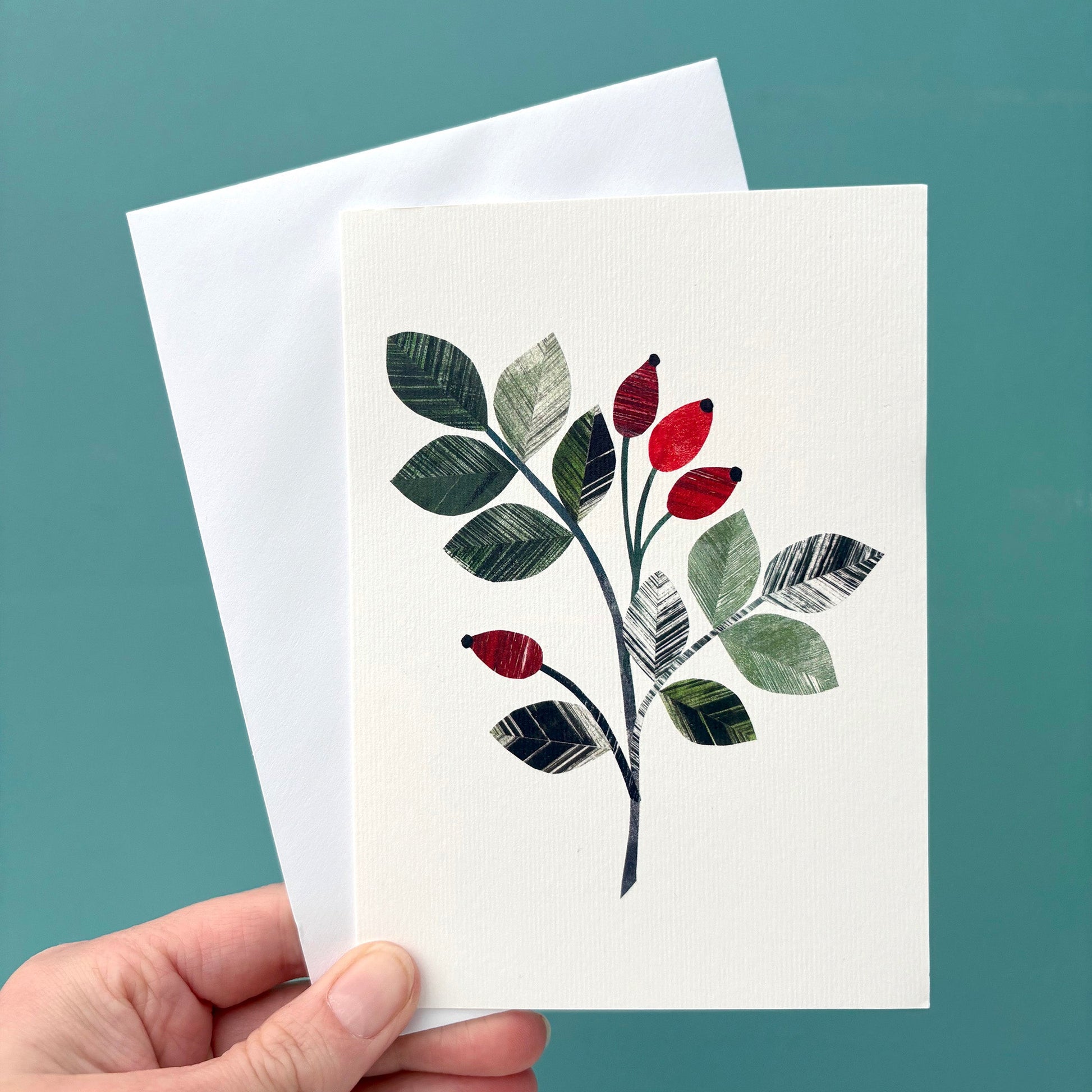 Hand holding a card with a Rosehip leaf sprig on it, featuring the bright red rose hip berries and textured green leaves.  It I being held with a white envelope against a teal background.