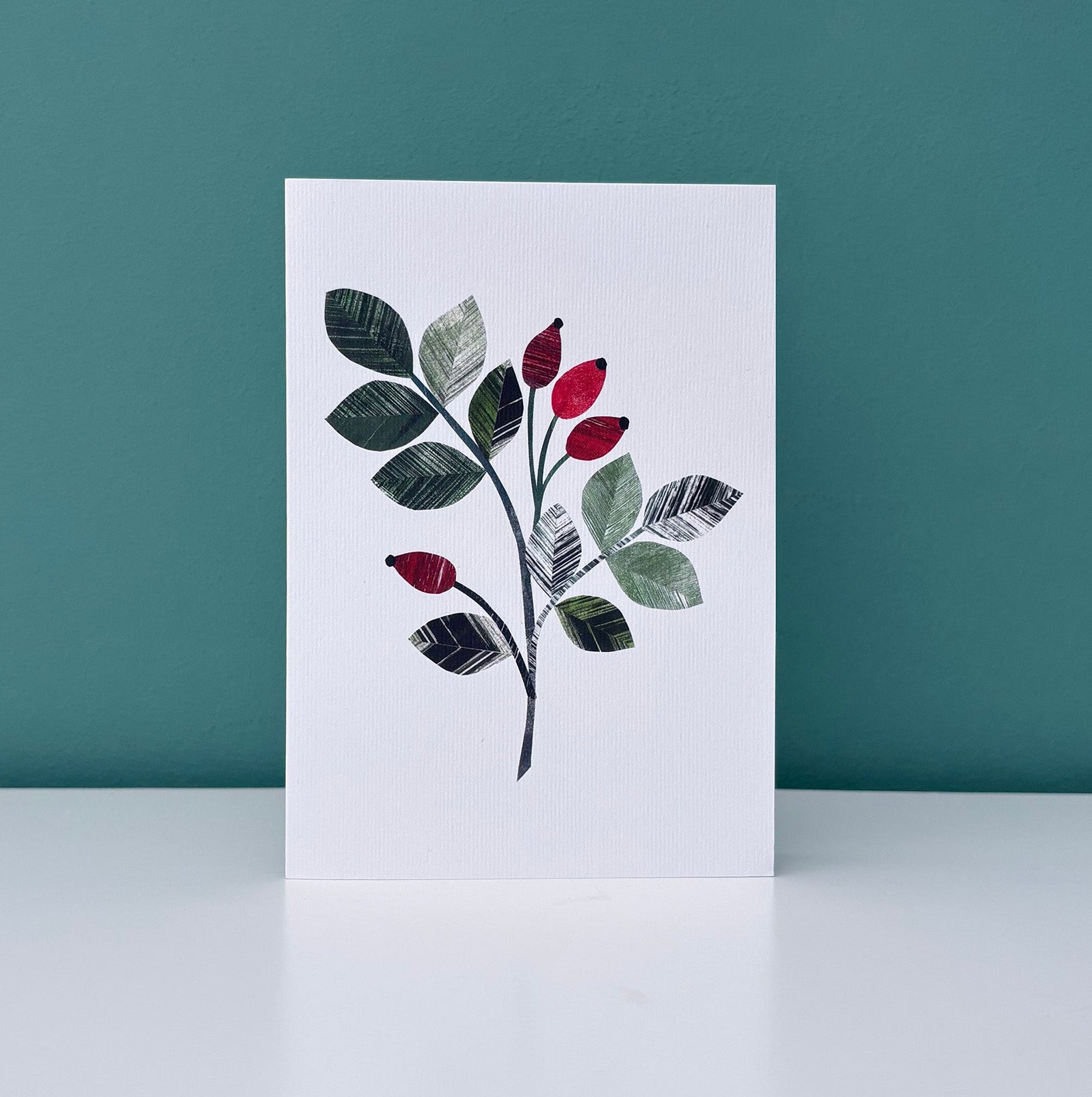 A card with a Rosehip leaf sprig on it, featuring the bright red rose hip berries and textured green leaves.  It has been placed on a white shelf with a Teal wall behind it.