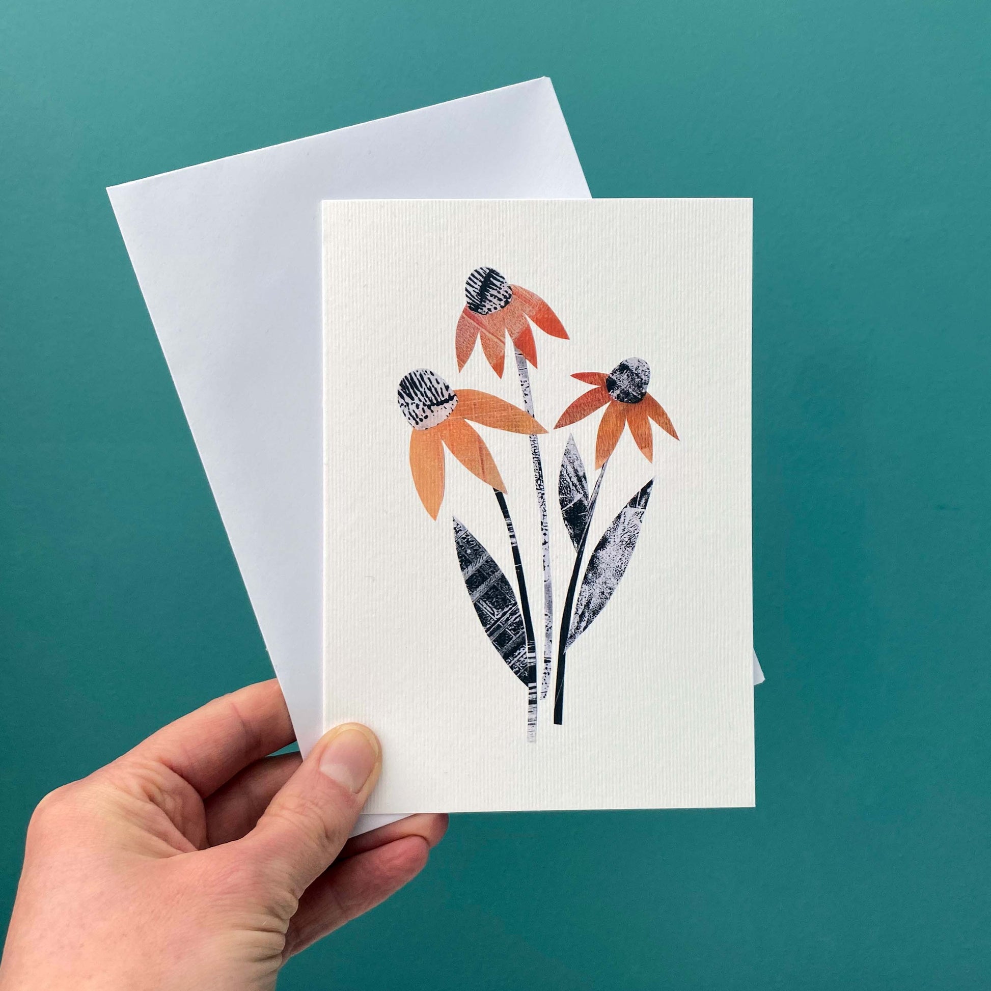 A Card with an orange rudbeckia flower on it one a white background is being held with a white envelope against a blue green wall.