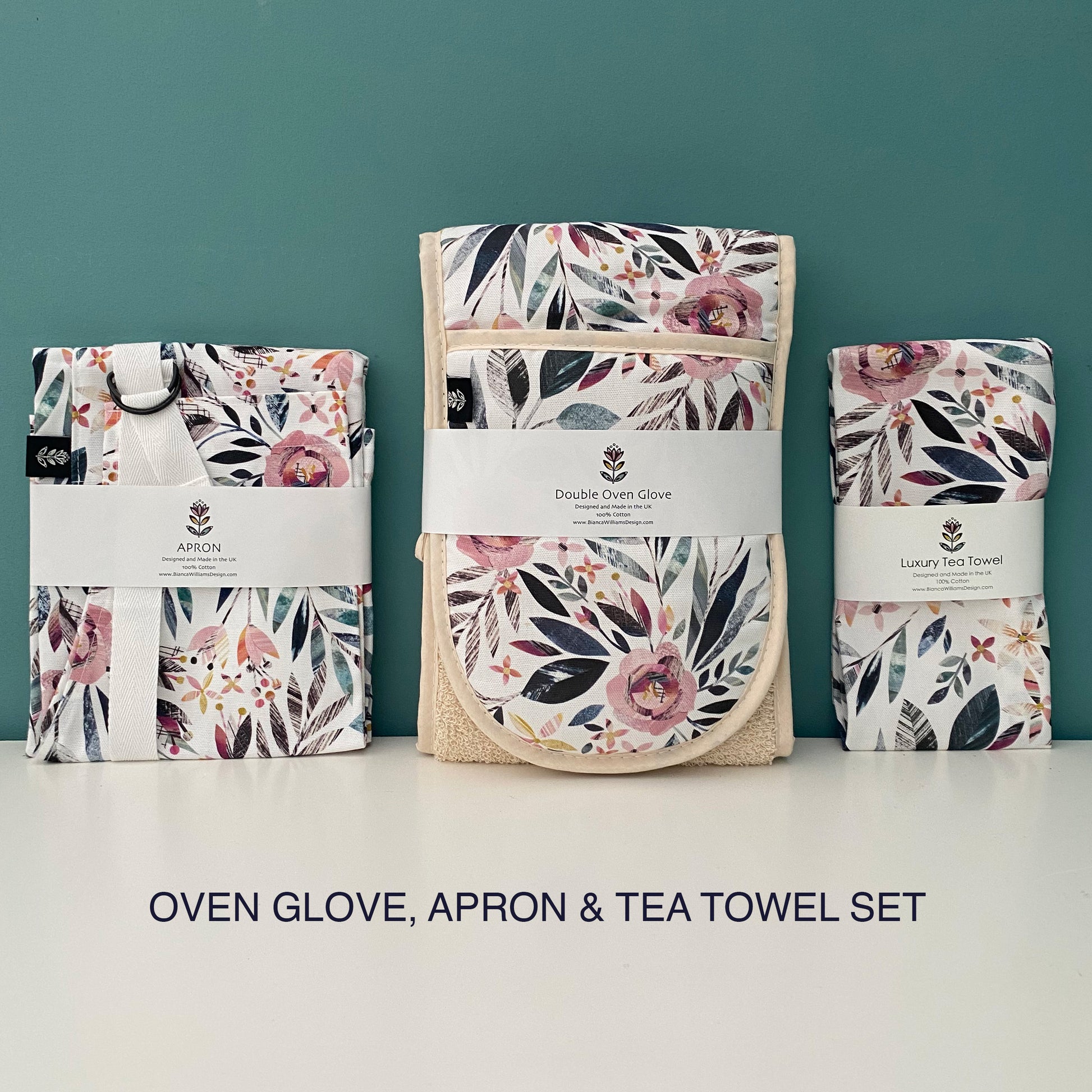 A packaged matching Summer Floral Apron, Double Oven Gloves and Tea Towel have all been placed on a white shelf with a blue green background.