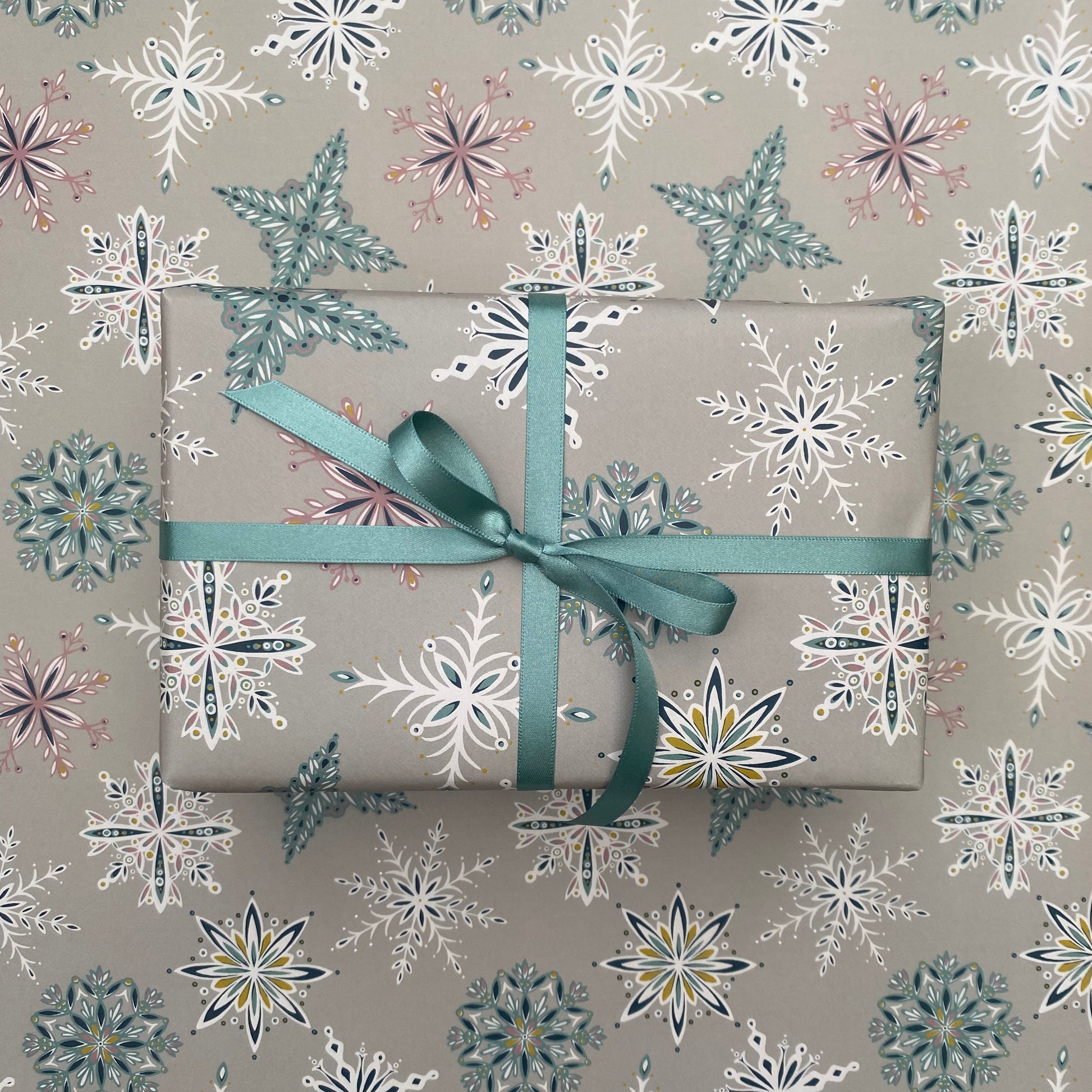 A present has been wrapped in the grey Snowflake gift wrap and has been tied with matching blue satin ribbon in a bow.  It has been placed on a sheet of the grey snowflake gift wrap.