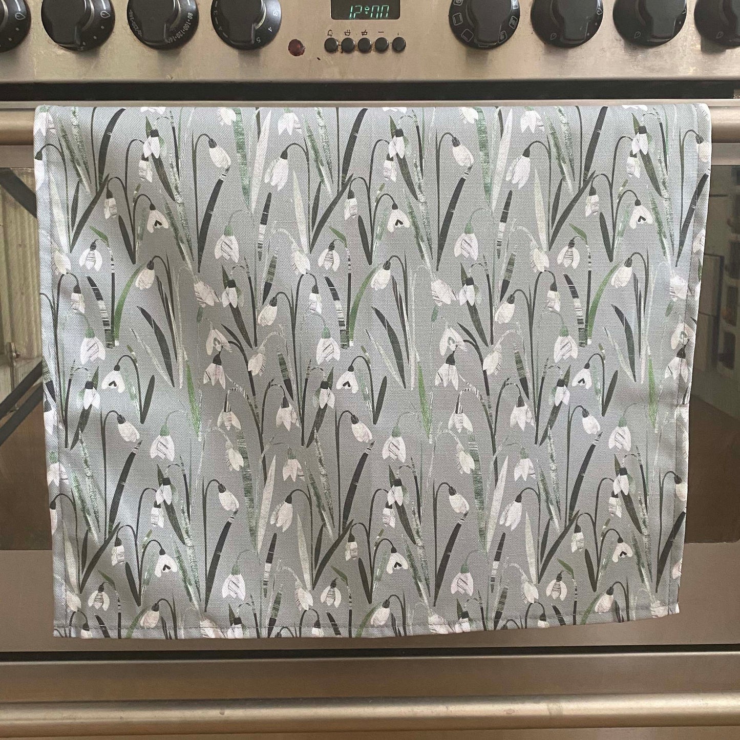 Snowdrops Tea Towel has been hung over the door of a stainless steel range cooker.