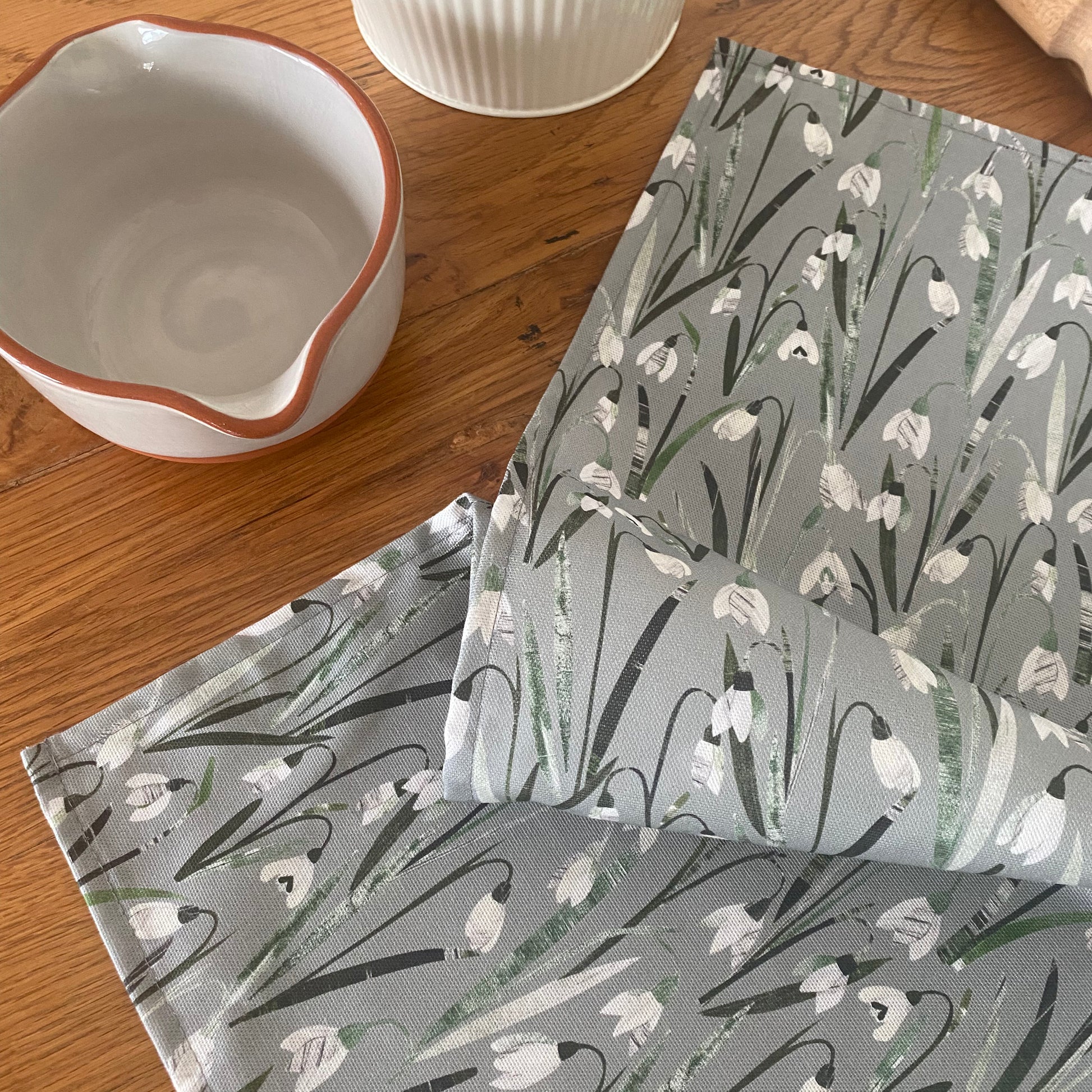 A snowdrops cotton tea towel featuring white and cream snowdrop flowers on a sage green background has been placed loosely on a Solid Oak Table.