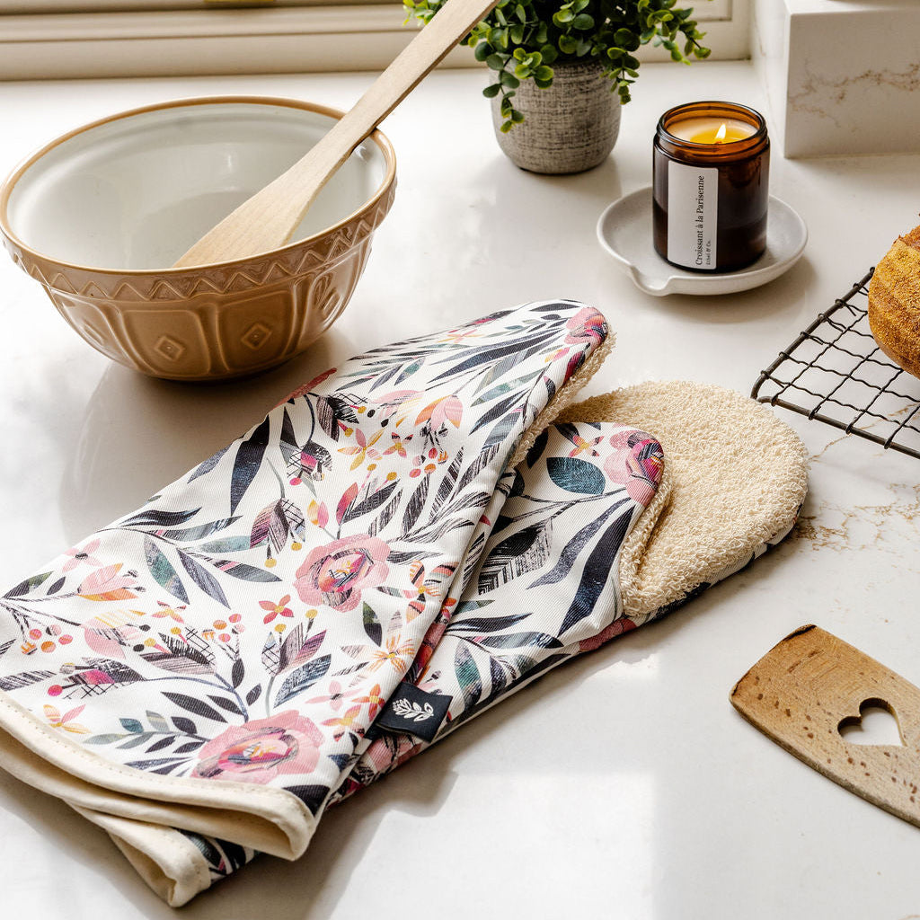 A pair of Summer Floral Long Oven Gloves have been placed on a marble kitchen worktop. They featuring a pink Roses pattern with green leaves on an off white background.  They have a beige cotton towel backing.  They have been placed on the worktop with a mixing bowel and a candle.