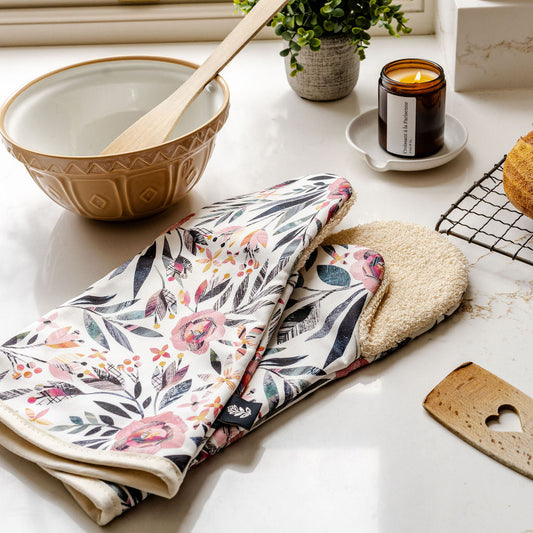 A pair of Summer Floral Long Oven Gloves have been placed on a marble kitchen worktop. They featuring a pink Roses pattern with green leaves on an off white background.  They have a beige cotton towel backing.  They have been placed on the worktop with a mixing bowel and a candle.