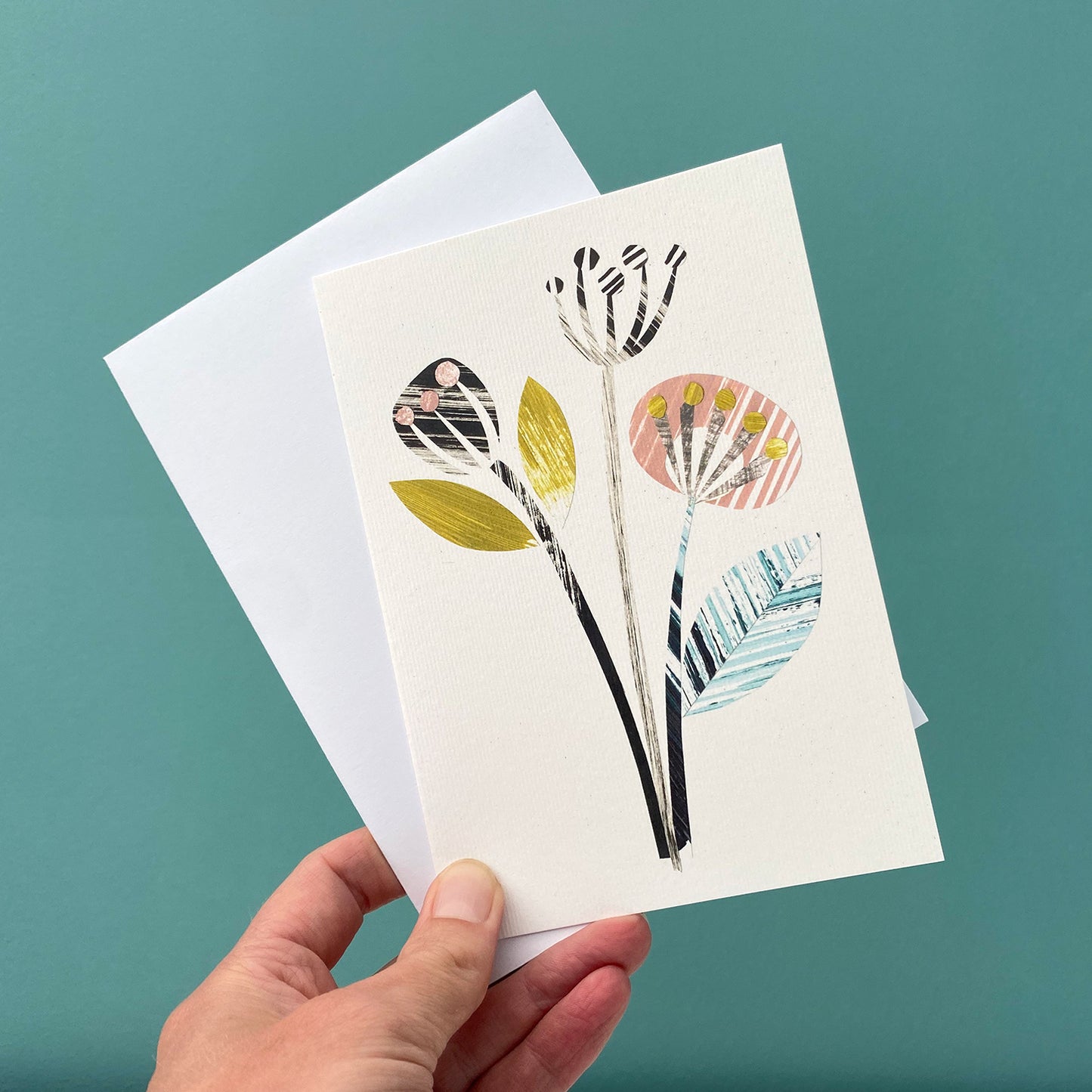 A card featuring a print of 3 stylised seed pods in yellow, green, pink and greys is being held with a white envelope against a blue green wall.