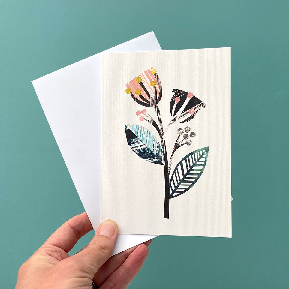 A card featuring a stylised version of 2 flower seed pods, in dusky pink, green and black and grey on a white back ground.  The card is being held with a white envelope against a blue green background.