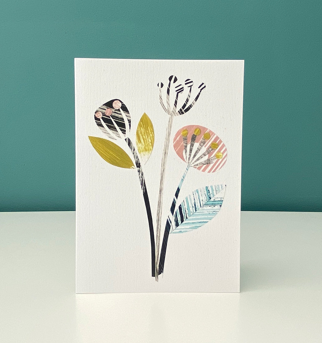 The card featuring a print of 3 stylised seed pods in yellow, green, pink and greys is stood on a white shelf with a blue green wall behind it.