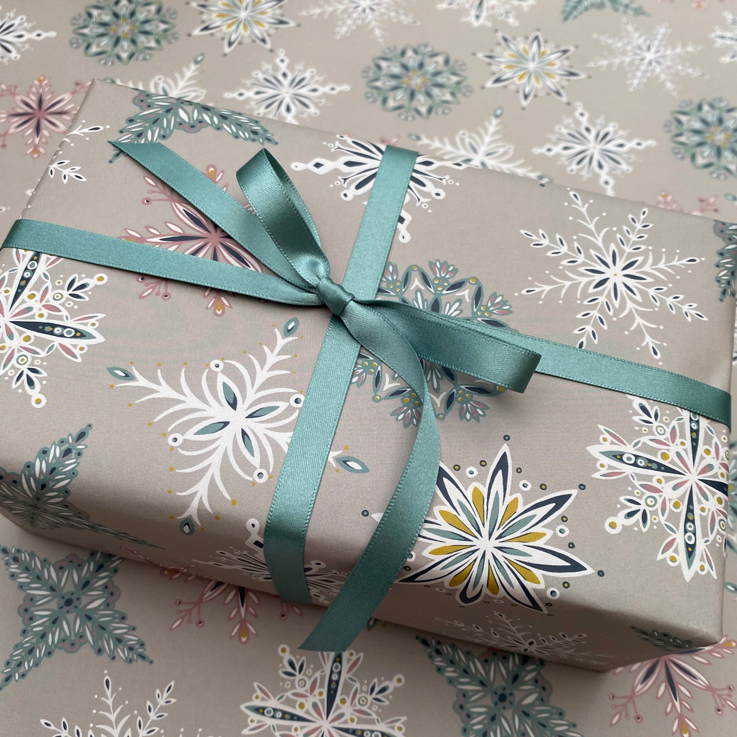 A close up of a present that has been wrapped in the grey Snowflake gift wrap and has been tied with matching blue satin ribbon in a bow.  It has been placed on a sheet of the grey snowflake gift wrap.