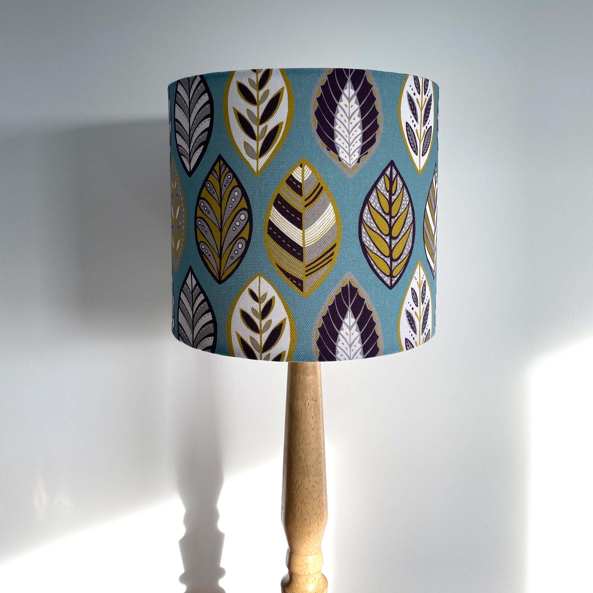 Small Blue Beech leaf Lampshade, shown here on a wooden table lamp. The pattern features a Skandi style Blue, yellow, grey and white Leaf pattern on a Blue Background.
