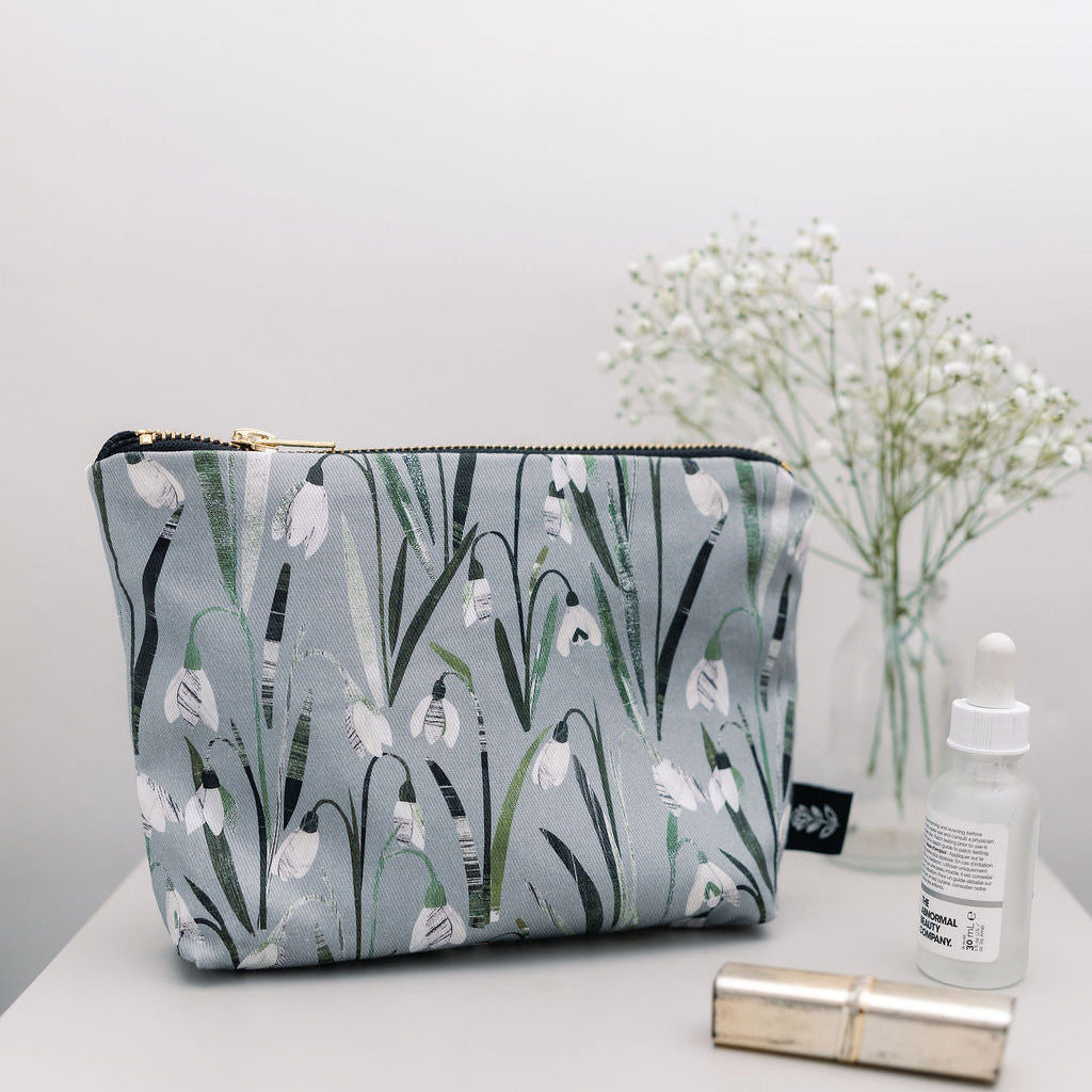 A rectangular zippered cosmetic pouch with a gray and white snowdrop floral print, styled on a white surface next to a small glass dropper bottle, a gold tube, and baby's breath flowers in a vase.