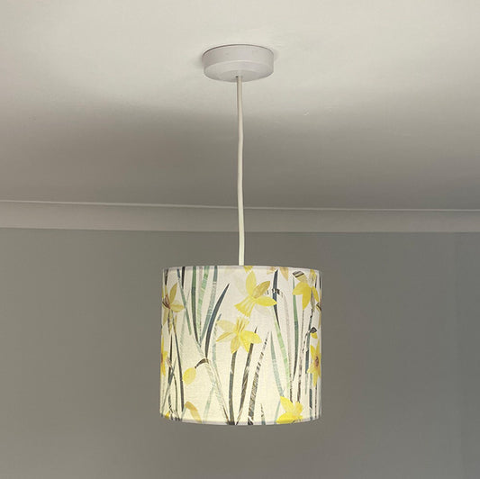 Daffodil Lampshade with a pendant ceiling fitting, in medium size. Has Yellow Daffodil flowers with Green Leaves on a plain white background. The lampshade is photographed against a blue/green background.  The light in on in this photograph.