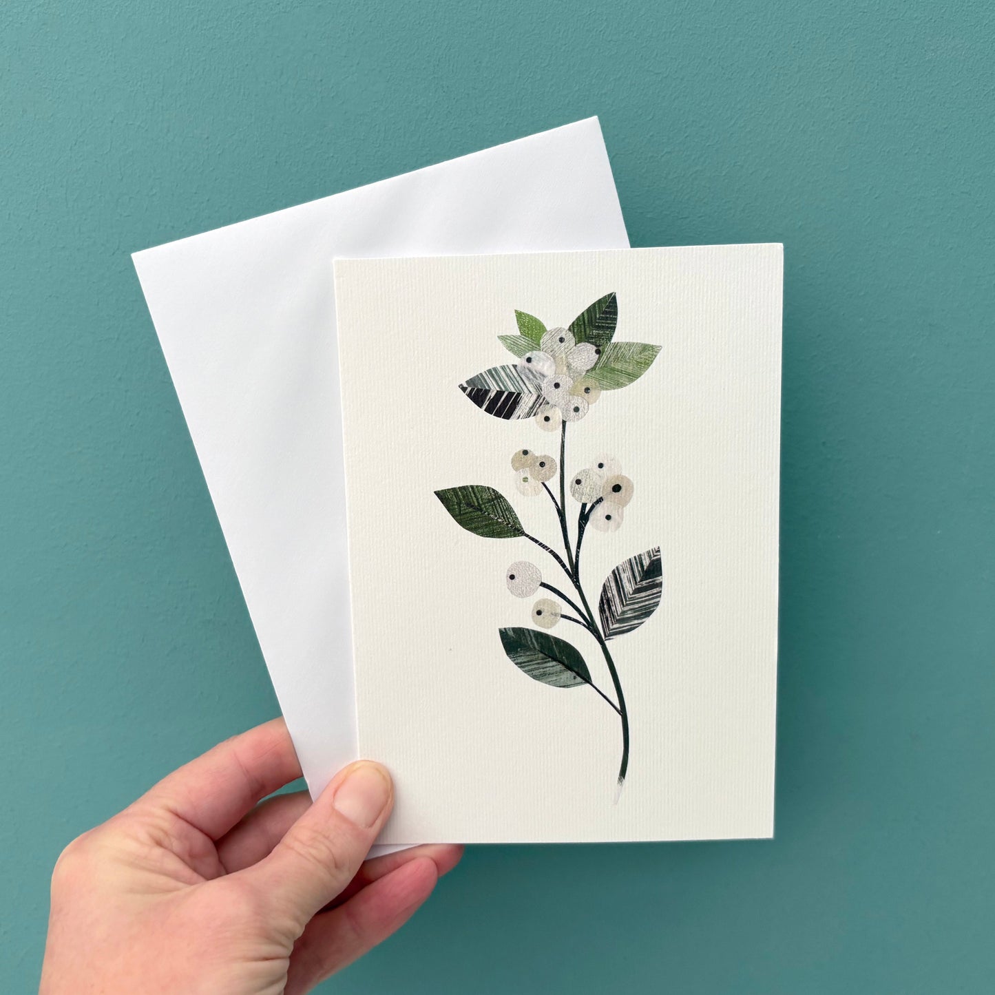 Hand holding a Greeting card with a winter floral design featuring a Snowberry sprig of delicate white berries with textured green leaves with a white envelope against a teal background.