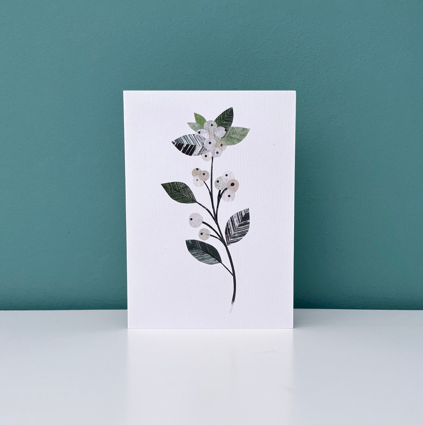 Greeting card with a winter floral design featuring a Snowberry sprig of delicate white berries with textured green leaves on a white background has been placed on a white shelf against a Teal wall.