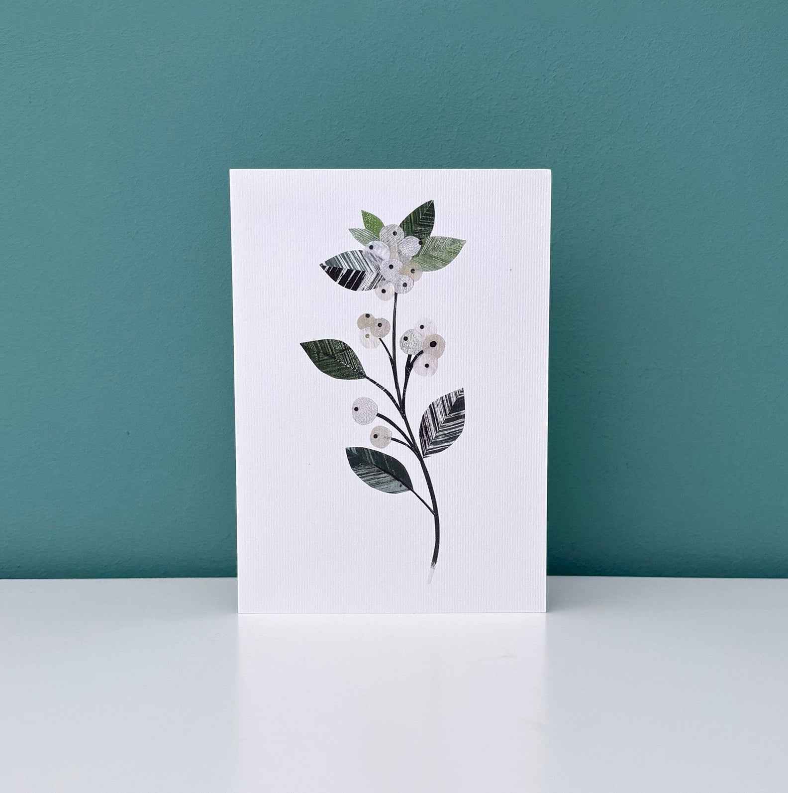 Greeting card with a winter floral design featuring a Snowberry sprig of delicate white berries with textured green leaves on a white background has been placed on a white shelf against a Teal wall.