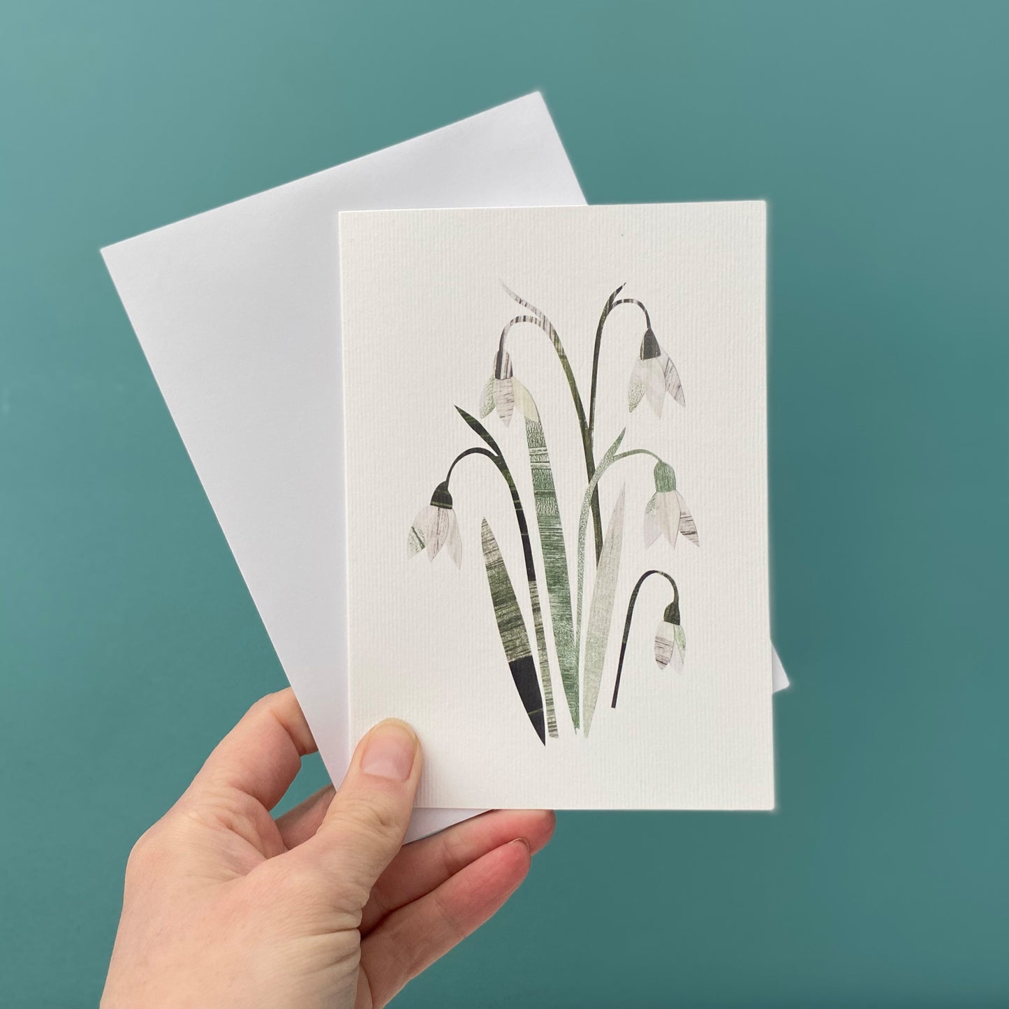 Snowdrop Greeting card featuring pretty cream and white snowdrop flowers with green leaves, is being held with a white envelope against a blue green background.