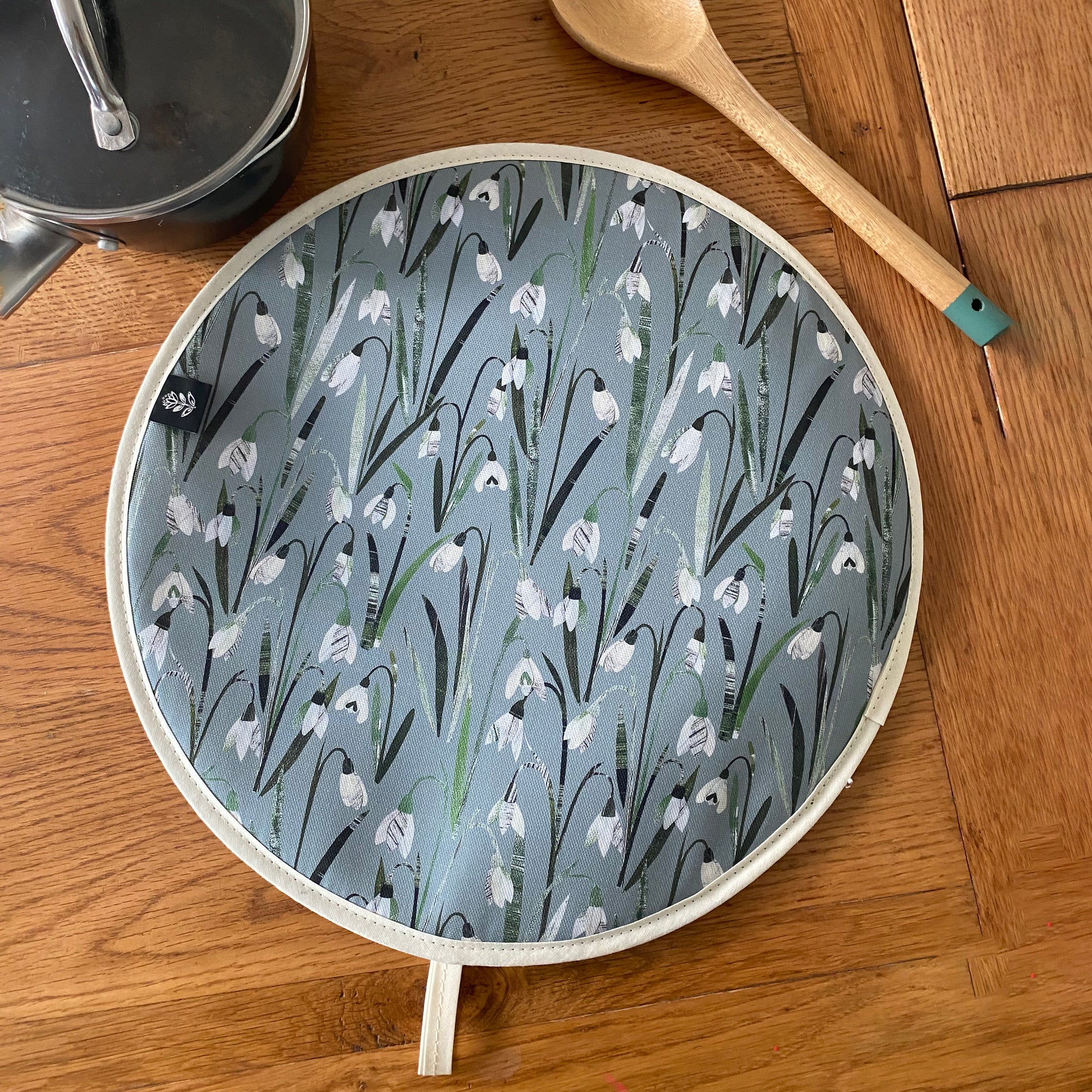 Snowdrops Aga Cover has been placed on a wooden table with a saucepan and a wooden spoon.