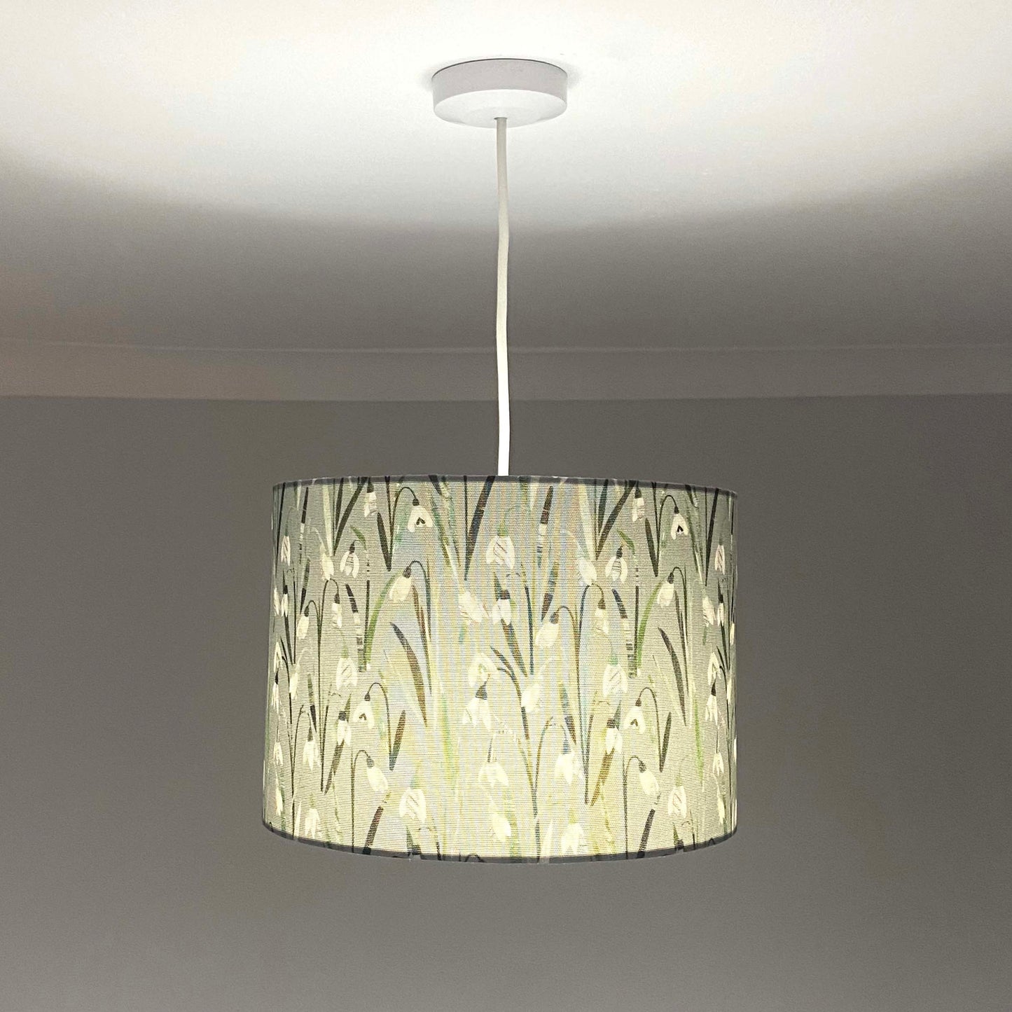 Snowdrop lampshades, shown here in a Size medium and as a Ceiling Pendant. The design features, white and cream snowdrop flowers with different shades of green leaves printed onto a pale grey/green background.  The light is switched on in this photograph.