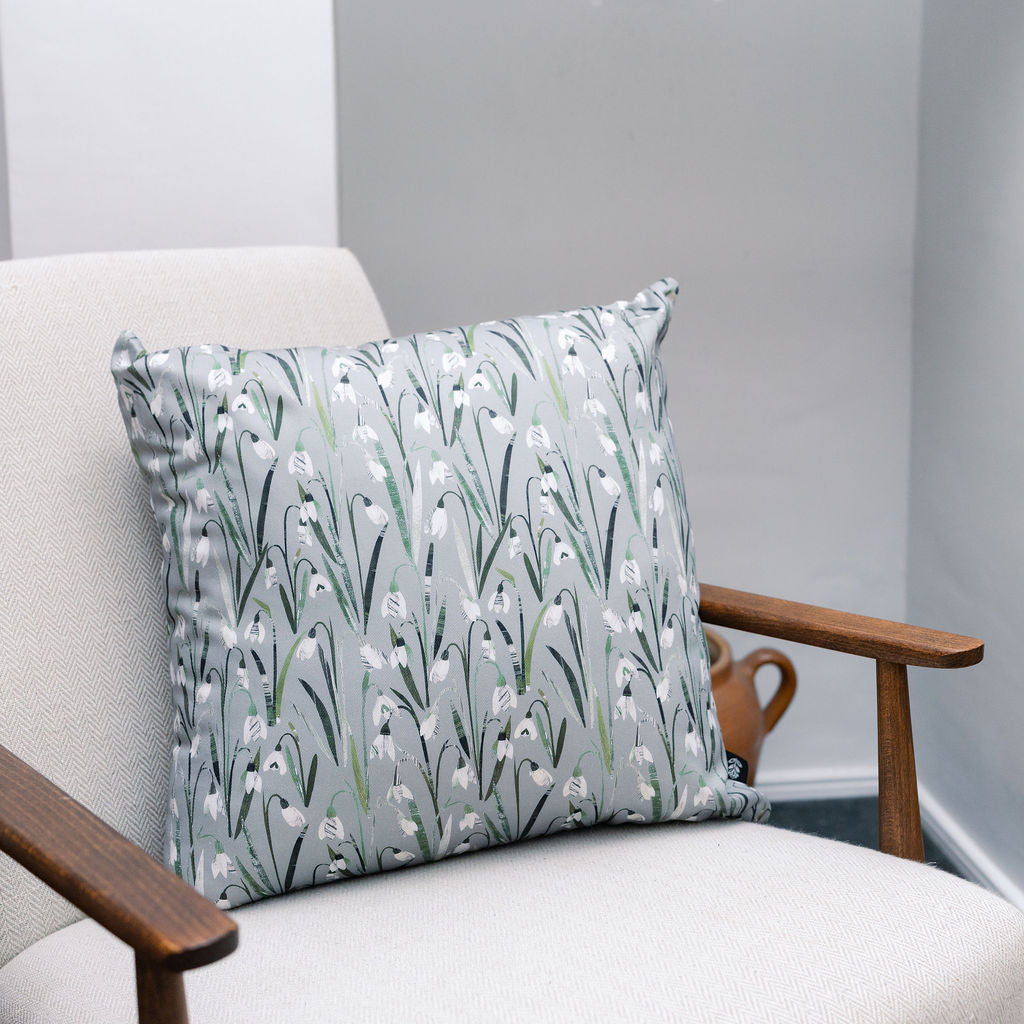 A square scatter cushion with a white snowdrop floral pattern on a muted green-grey background, resting on a light beige upholstered armchair with dark wood arms.
