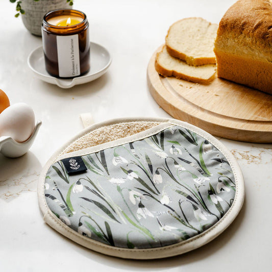 A round fabric oven mitt with a snowdrop pattern on a soft green grey background with a beige trim, resting on a white countertop next to a loaf of bread, sliced bread on a wooden board, a lit candle, and a small plant.