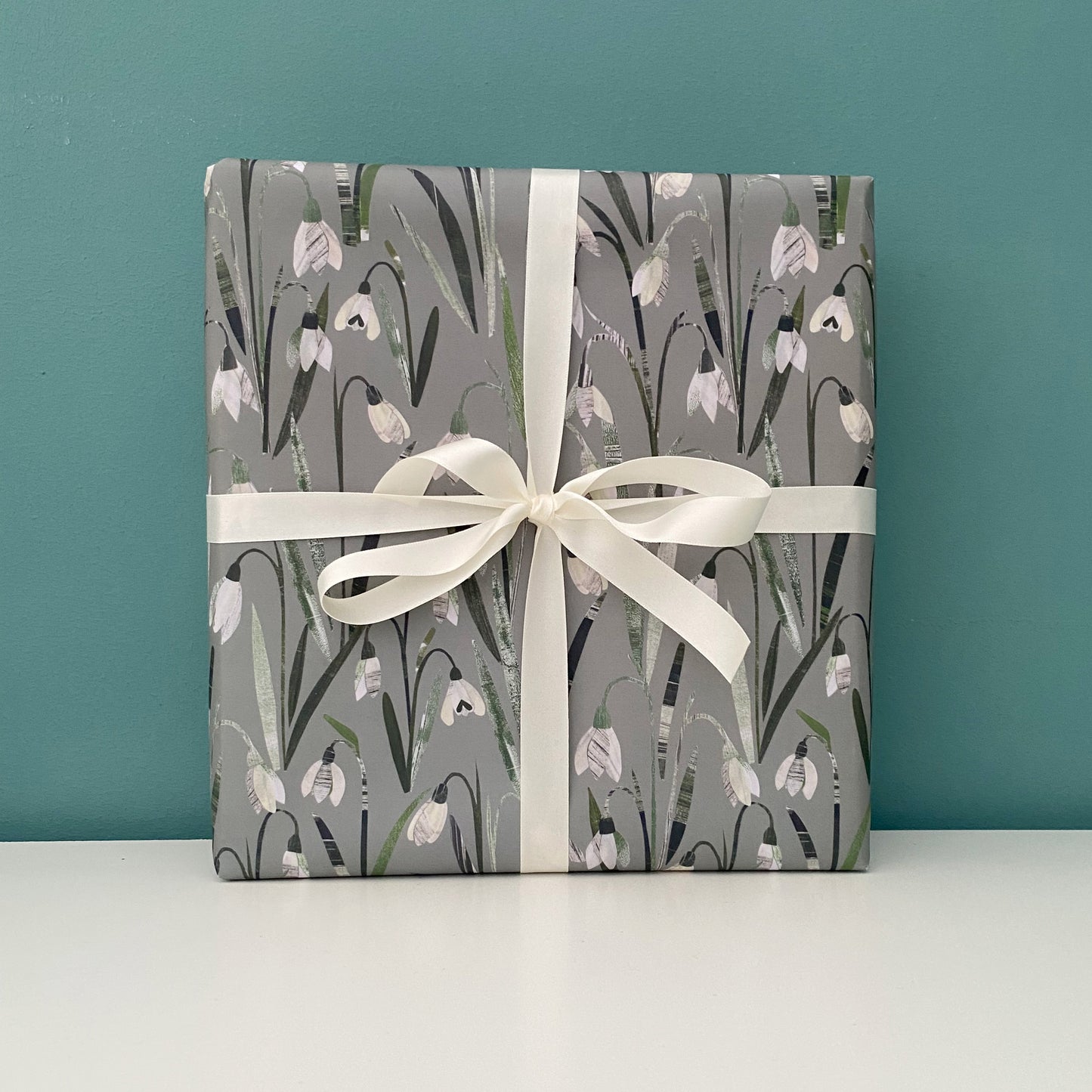 A present wrapped in Snowdrop gift wrap and ties with a cream satin bow has been placed on a white shelf against a blue/green wall.