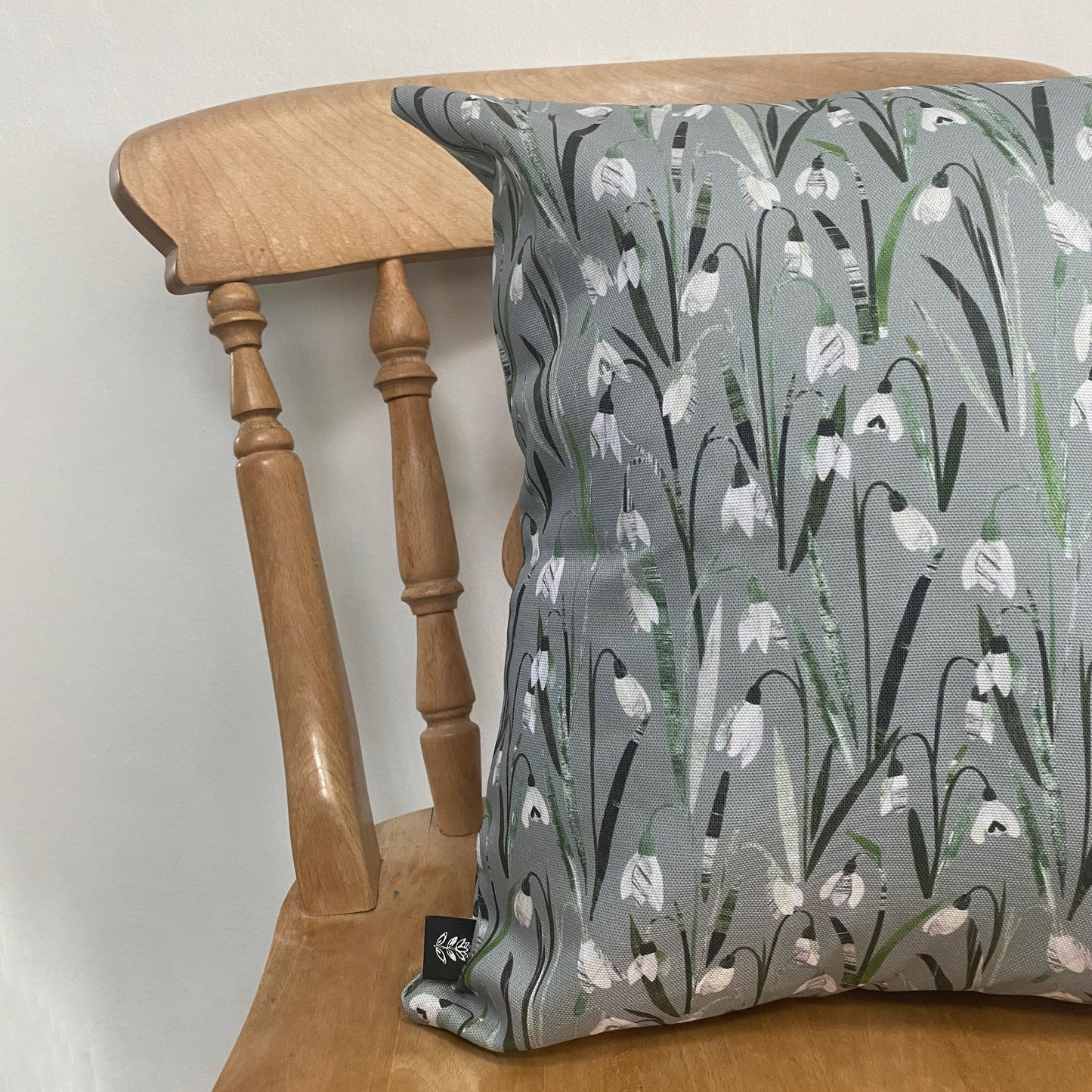 A Snowdrop Cushion has been placed on a wooden dining chair in front of a pale grey wall.