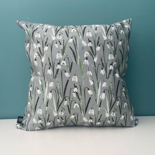 A Snowdrop Cushion featuring pretty little cream and white snowdrop flowers with green leaves on a soft sage green background.  The cushion has been placed on a white shelf against a blue/green wall.