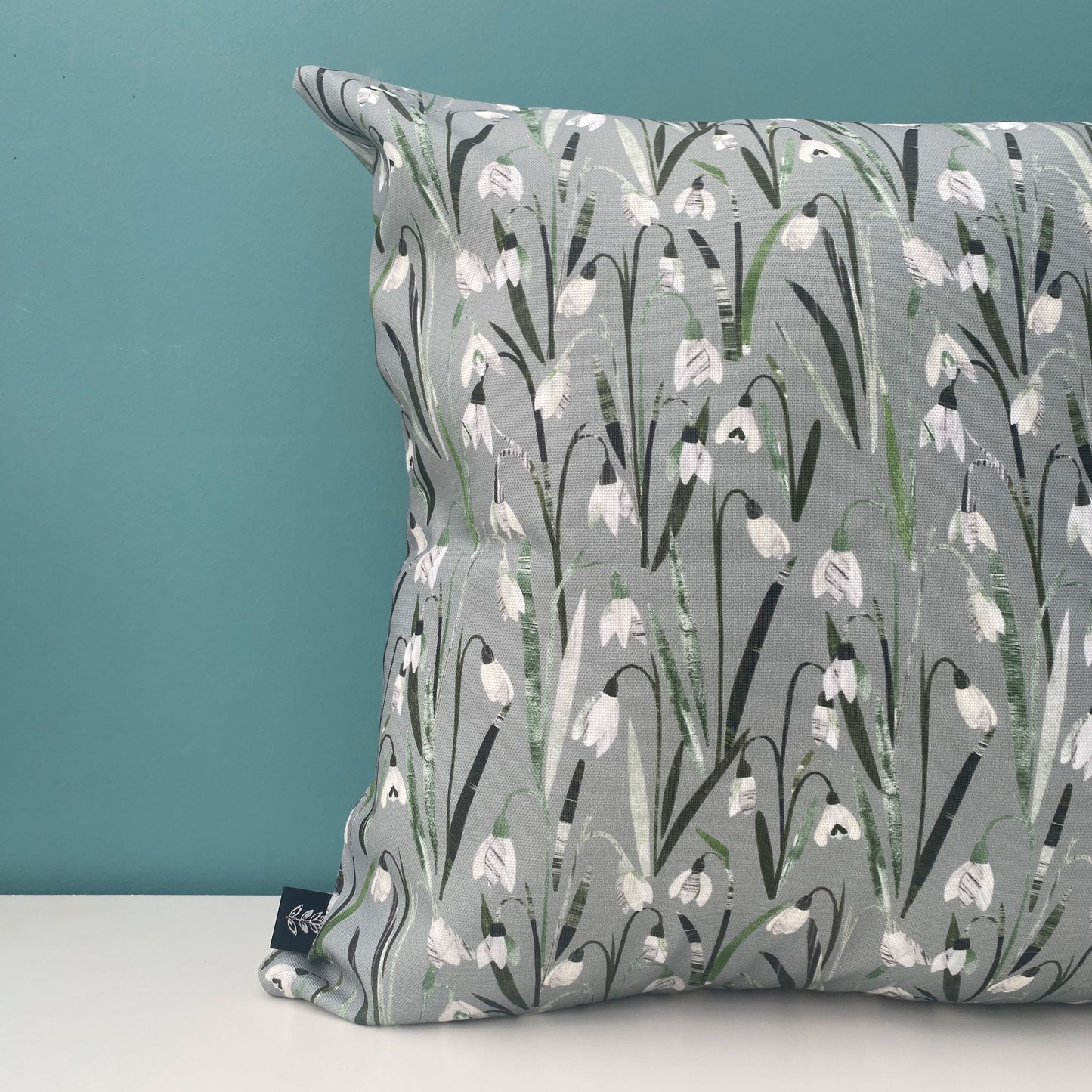 A close up of a Snowdrop Cushion featuring pretty little cream and white snowdrop flowers with green leaves on a soft sage green background. The cushion has been placed on a white shelf against a blue/green wall.