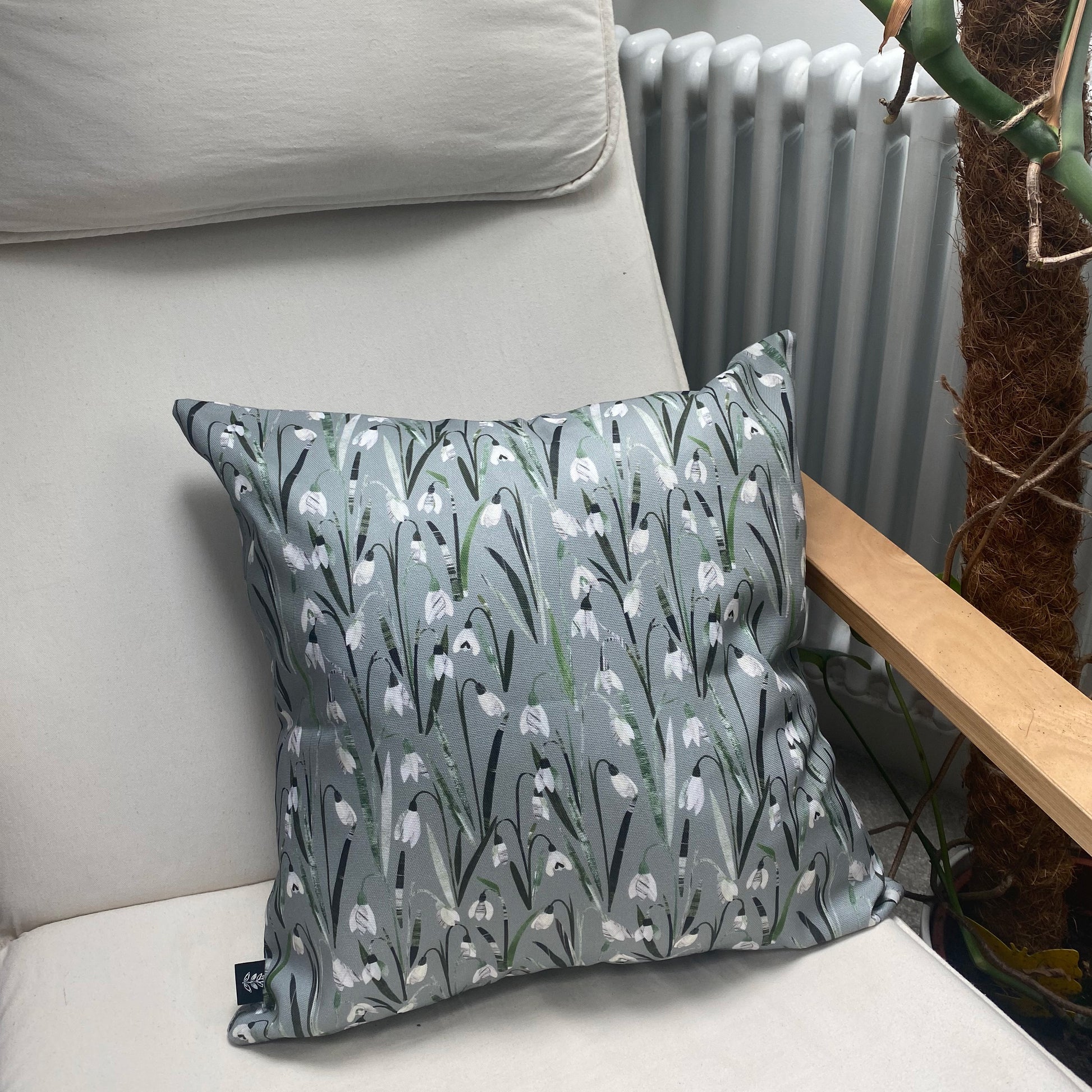 A Snowdrop Cushion has been placed on a cream canvas and wooden arm chair.  A radiator and plant can be seen in the background.