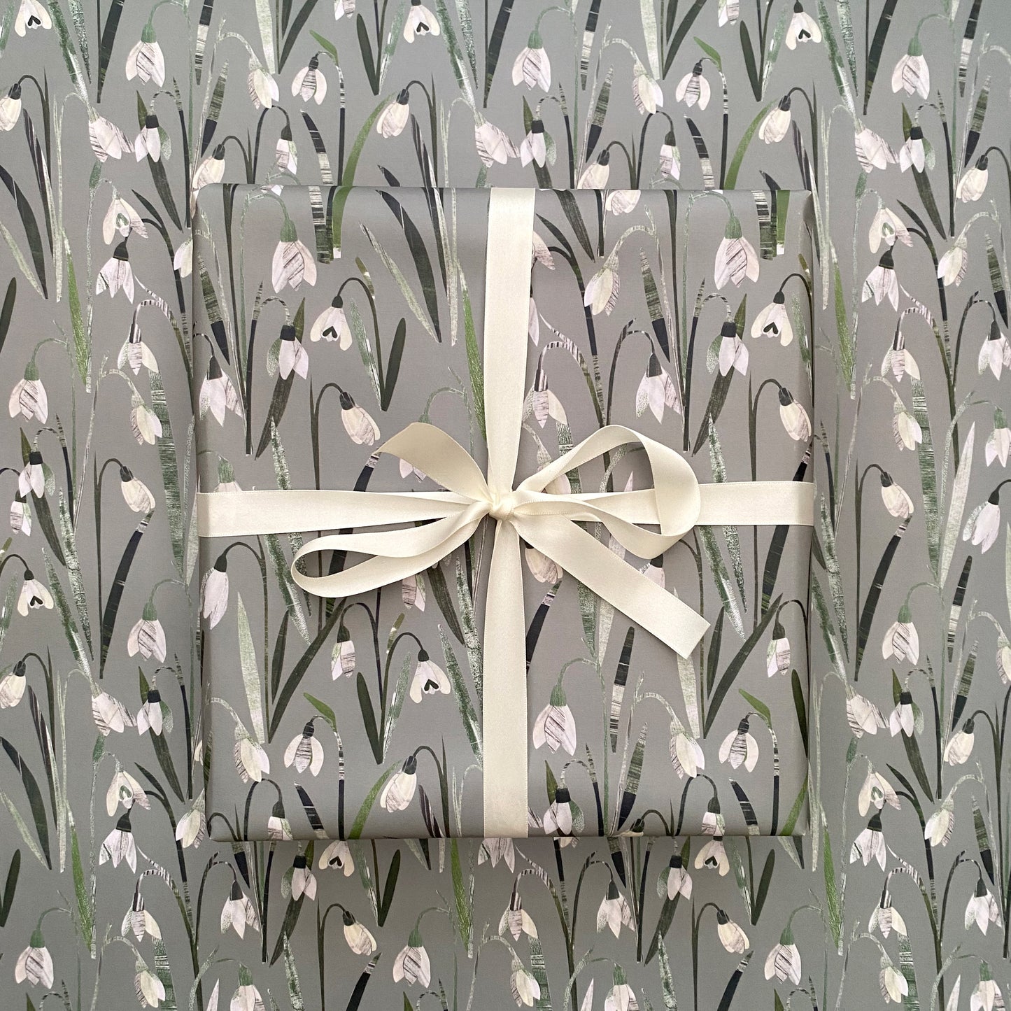 A present has been wrapped in Snowdrop Gift Wrap and has been tied with a cream satin bow.  The present has been placed on a sheet of gift wrap feating the same snowdrop design.