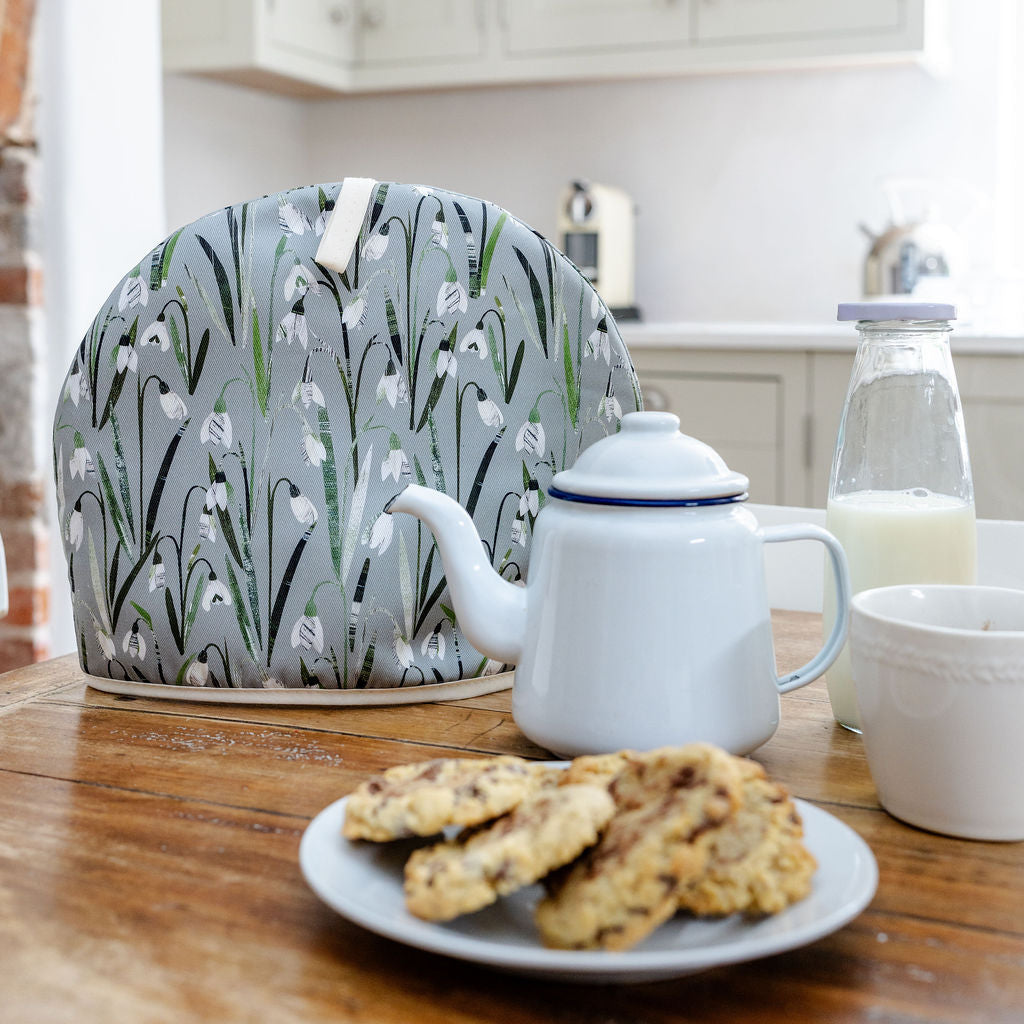  Snowdrops Tea Cosy has been placed on a wooden table with a plate of cookies a kettle and some milk.
