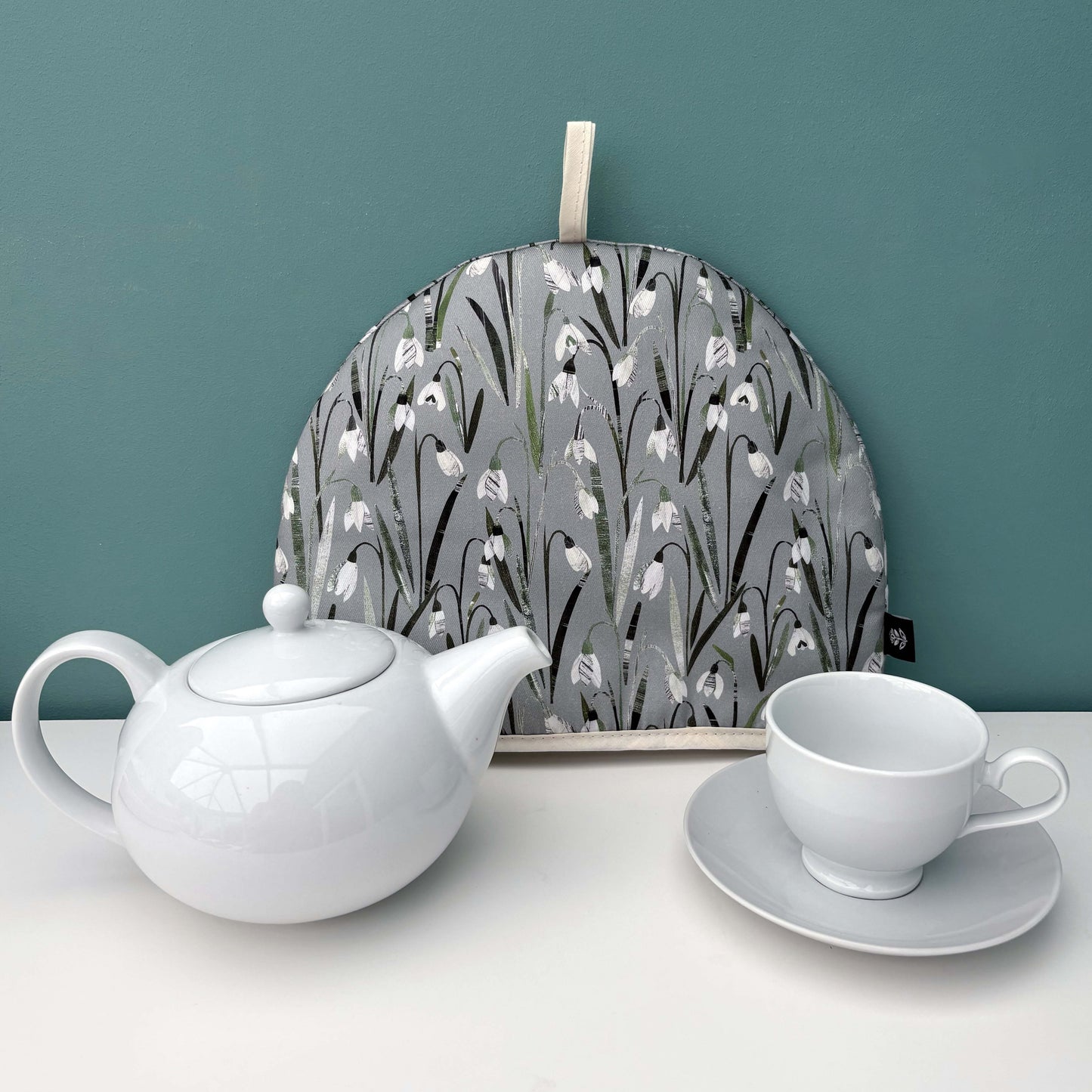 A Snowdrop Tea Cosy and a white teapot, cup, and saucer on a white surface against a teal wall.
