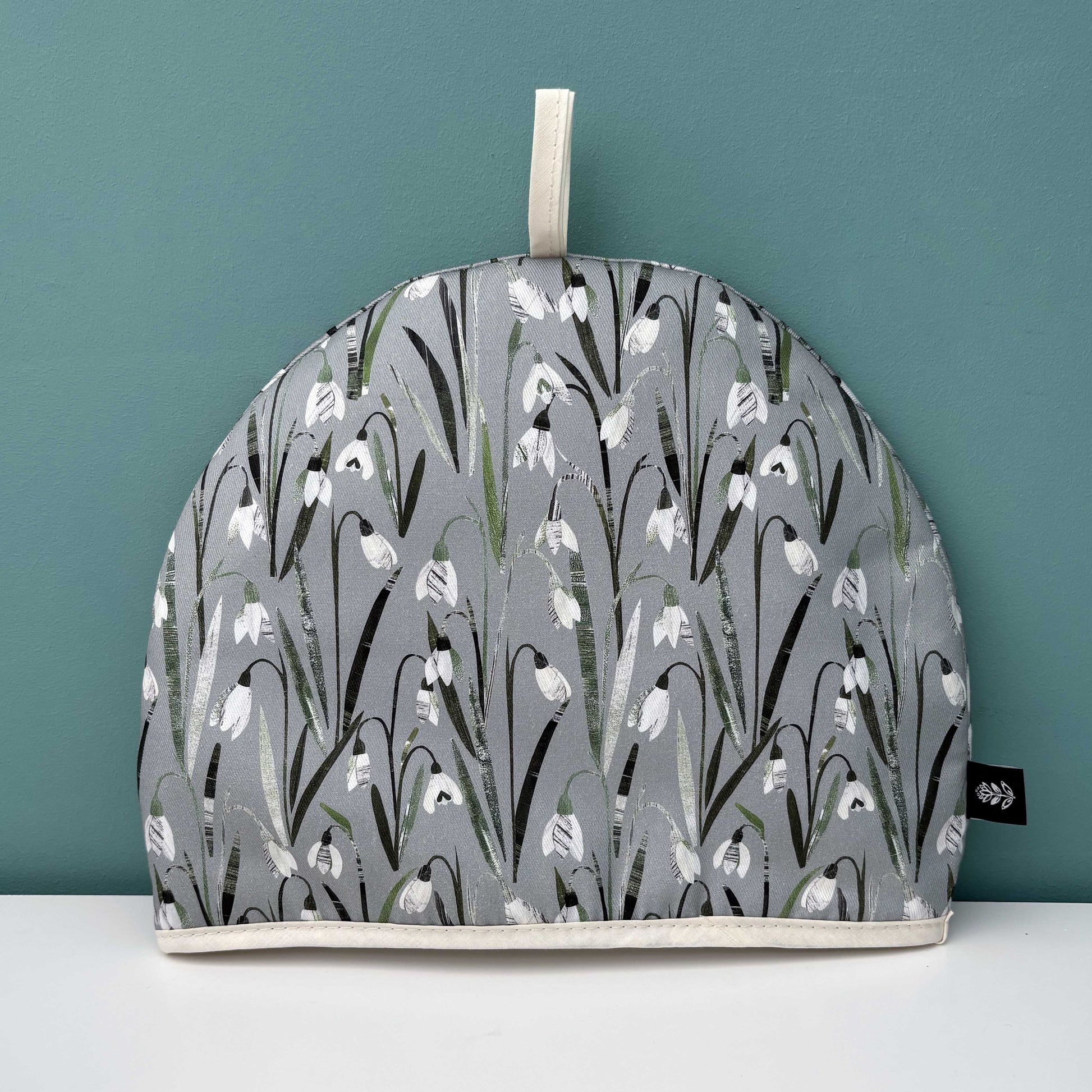 A Snowdrop Tea cosy with a white and cream floral pattern on a soft sage green background has been put on a white surface against a teal wall.
