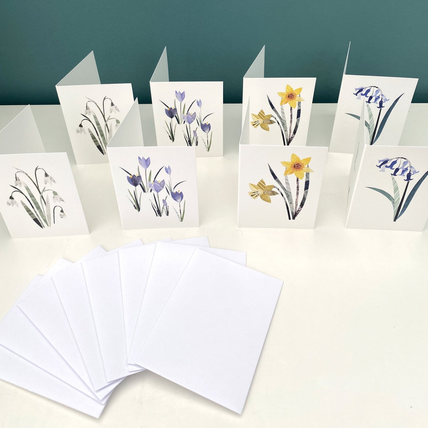 8 notecards featuring Daffodils, Crocus, Snowdrops and Bluebells are standing on a white shelf, 8 plain white envelopes are next to them.