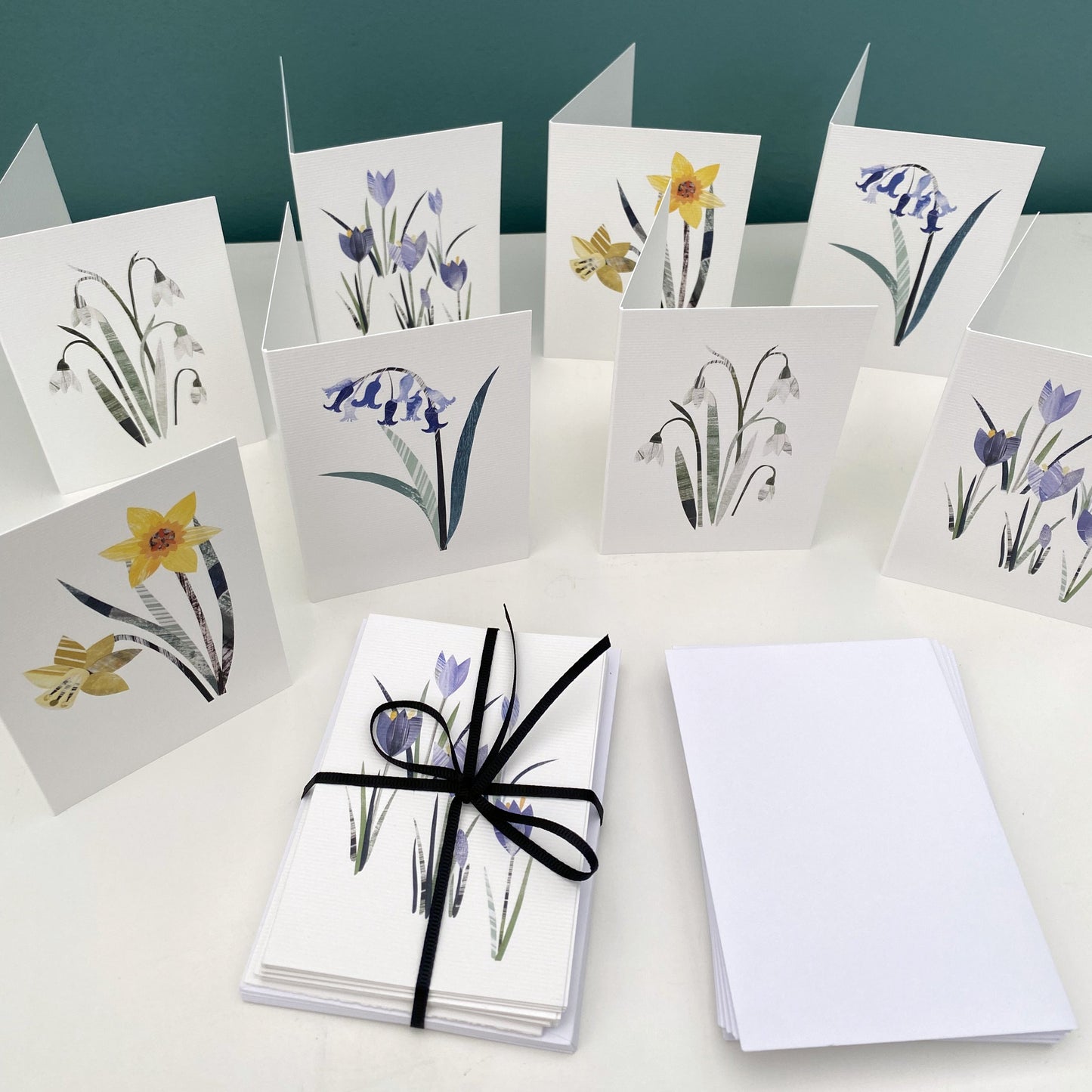 A pack of Spring Floral Note Cards have been tied with black ribbon and placed on a white shelf next to some white envelopes and the 8 cards in the pack have been stood behind on the shelf.