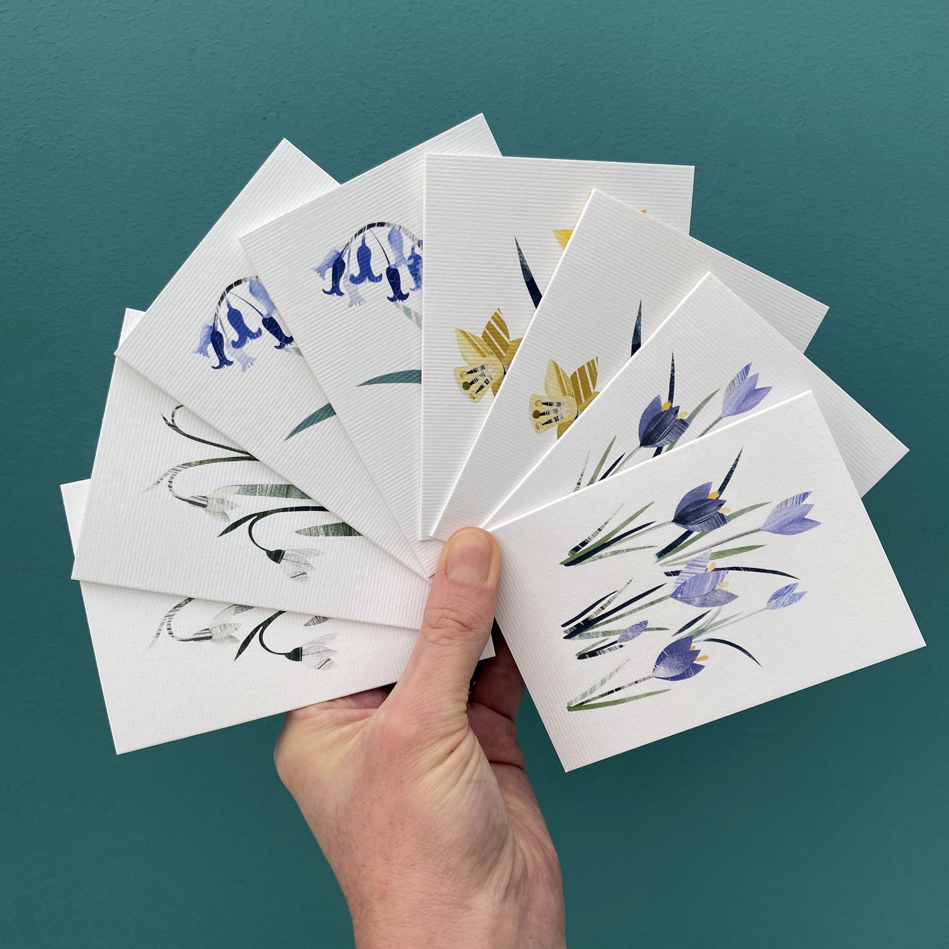 8 mini notecards featuring Bluebells, Snowdrops, Daffodils and Crocus are being held by hand in front of a blue green background.