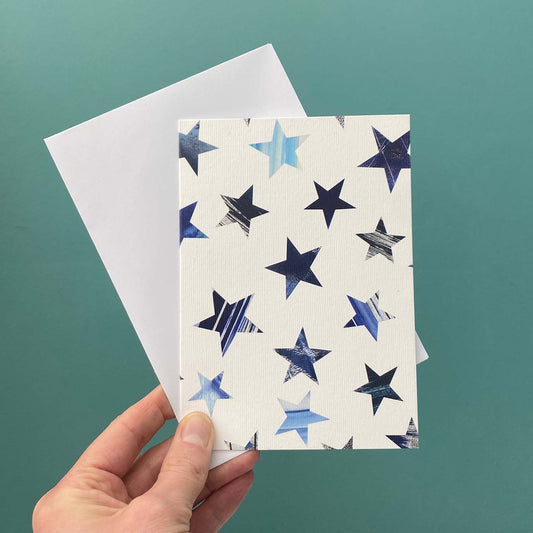 A hand holds a white textured greeting card with a pattern of blue, navy, and silver textured stars, with a plain white envelope behind it against a teal background.