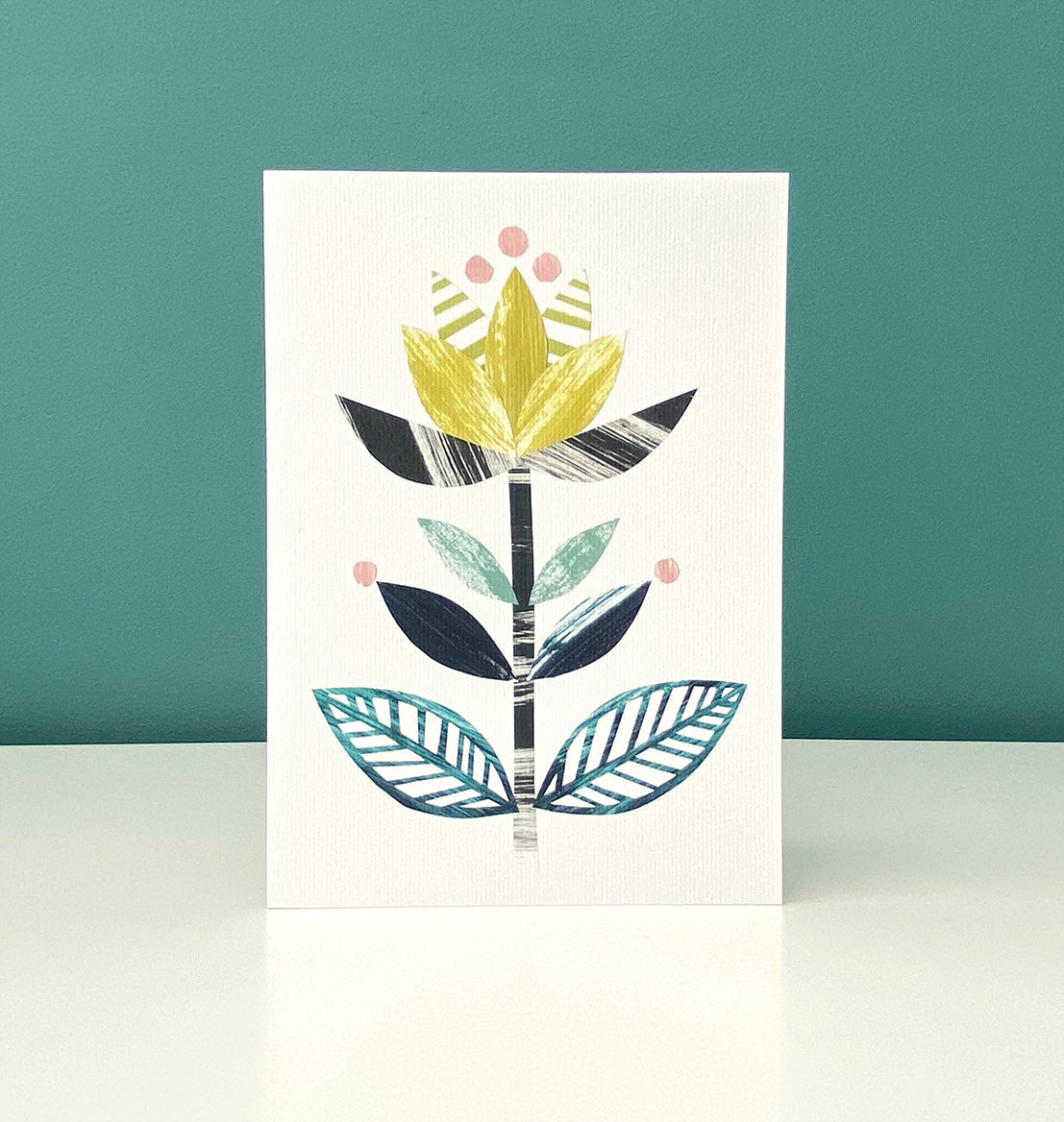 A card featuring a stylised flower motif in yellow, greens and blacks on a white background has been stood up on a white shelf with a blue green background.