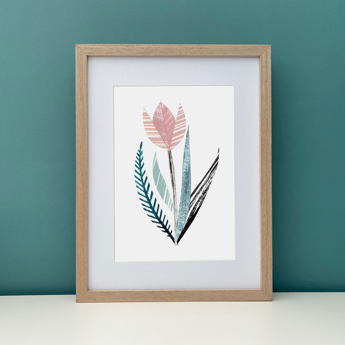 The stylised Pink Tulip Print has been put in a wooden frame and is on a white shelf against a teal wall.